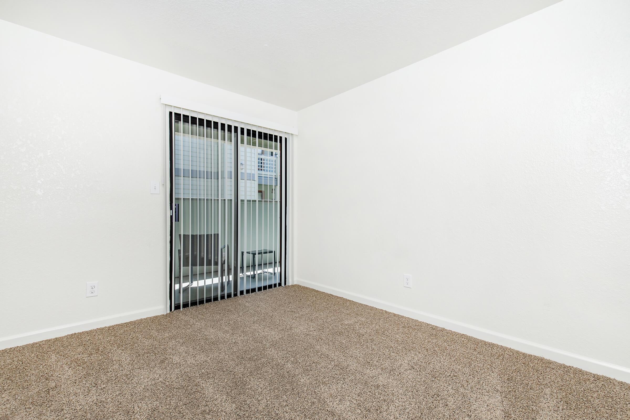 Empty room with beige carpet and white walls. A sliding glass door with vertical blinds leads to a balcony, allowing natural light to enter. The room is minimally decorated, creating a spacious feel. No furniture is present, emphasizing the openness of the space.