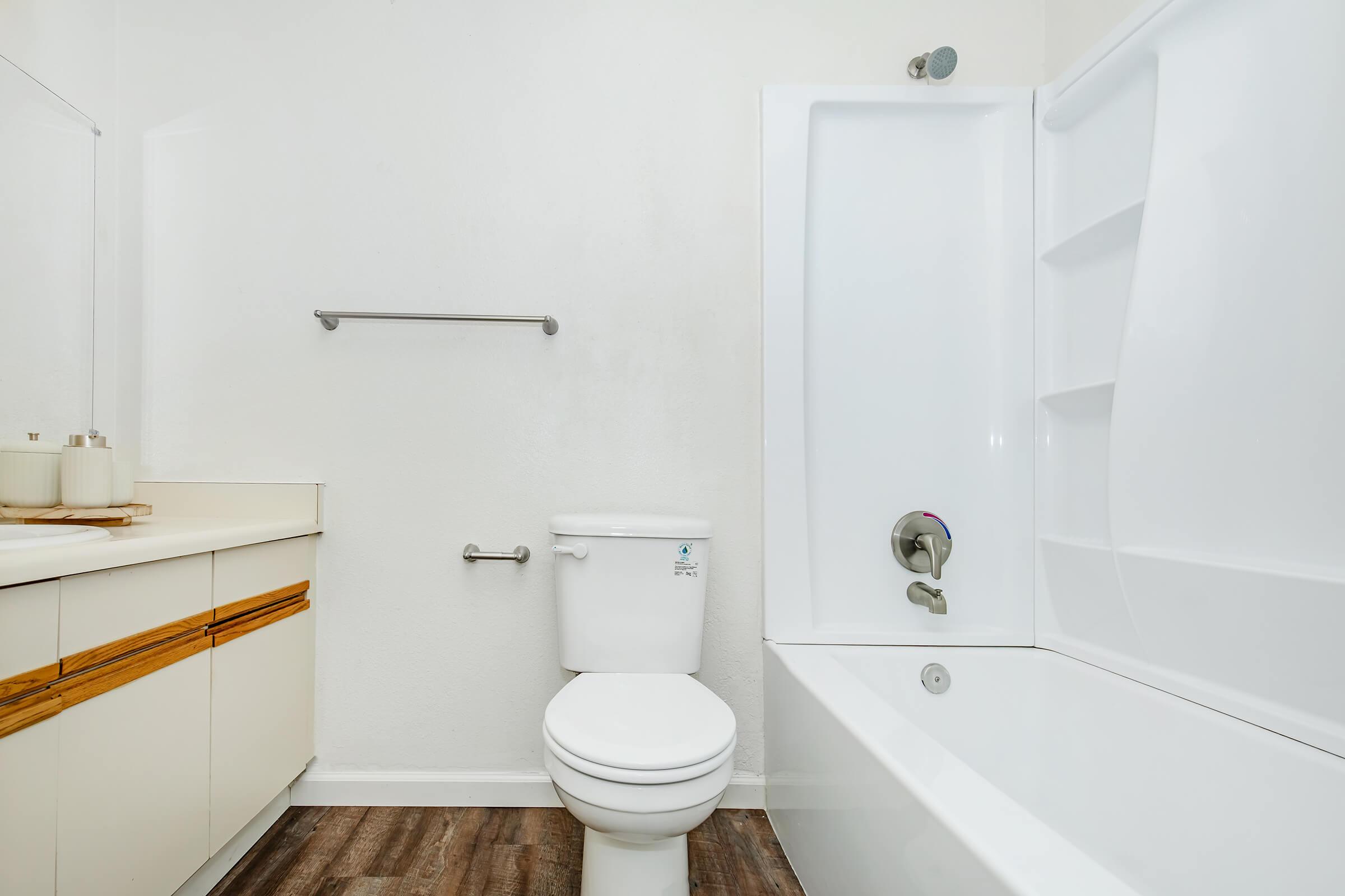 A clean and modern bathroom featuring a white bathtub with a shower, a white toilet, and a sink with a wooden cabinet below. The walls are light-colored, and there is a towel bar beside the toilet. The flooring has a wood-like appearance, contributing to a simple and minimalist aesthetic.