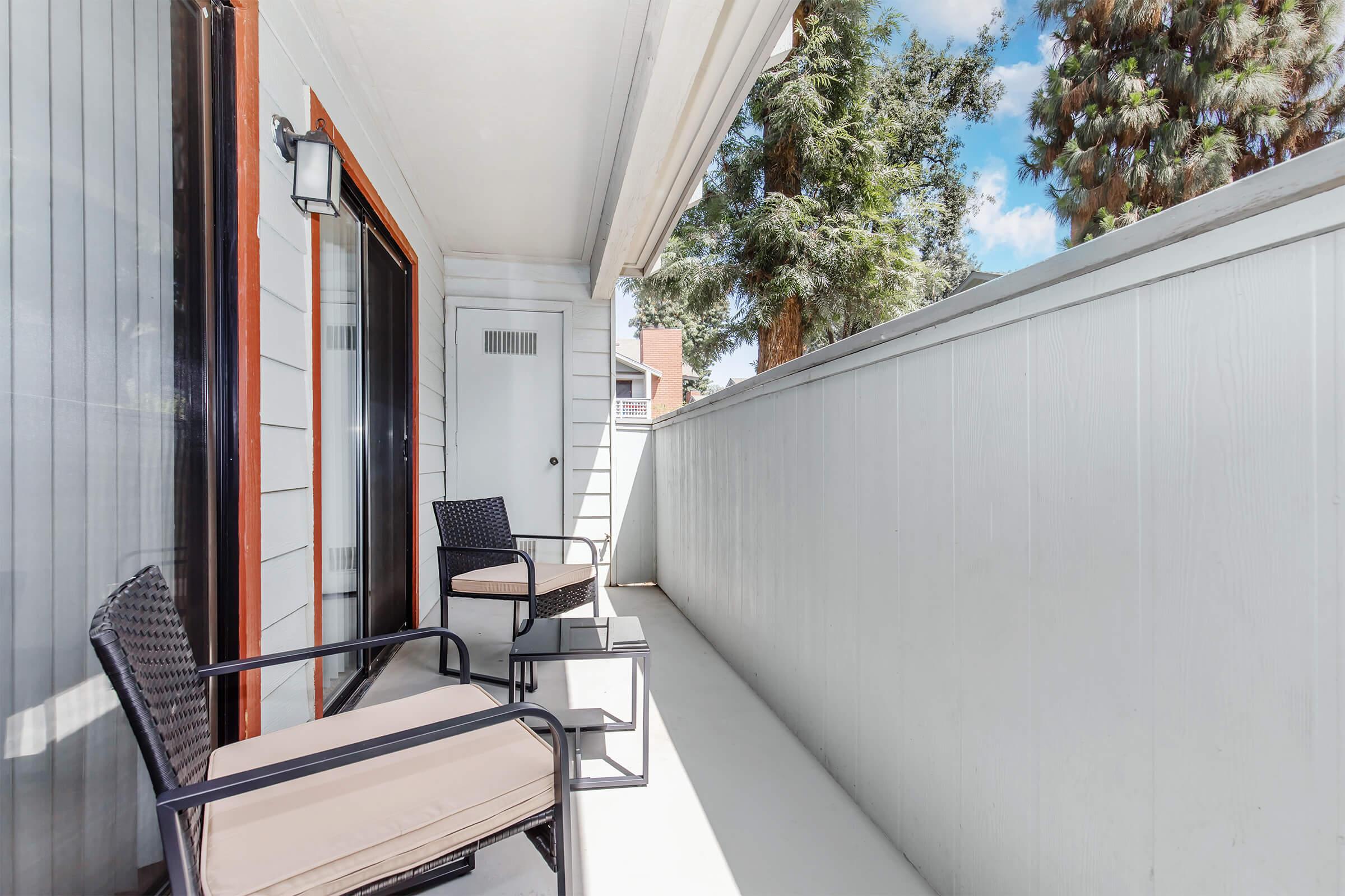 A small balcony with two wicker chairs and a side table, surrounded by tall trees. The walls are painted white, and there are sliding glass doors leading inside. The sky is blue with some clouds, creating a bright and inviting outdoor space.
