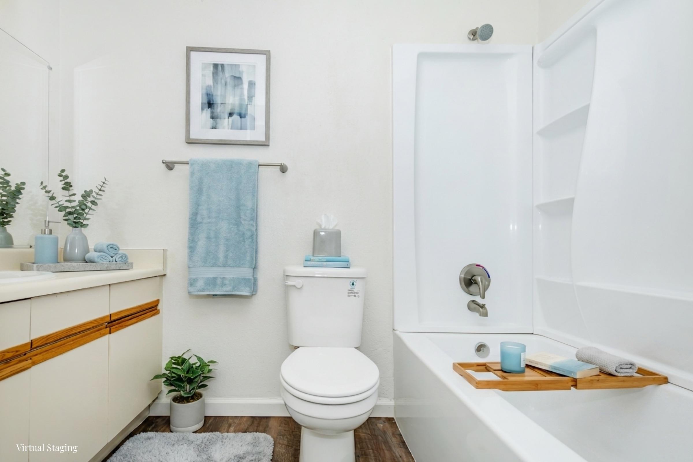 A clean and bright bathroom featuring a white bathtub with a wooden tray, a white toilet, and a vanity with a mirror. Light blue towels hang on a towel bar, and a decorative plant adds a touch of greenery. A piece of artwork is displayed on the wall, enhancing the aesthetic.
