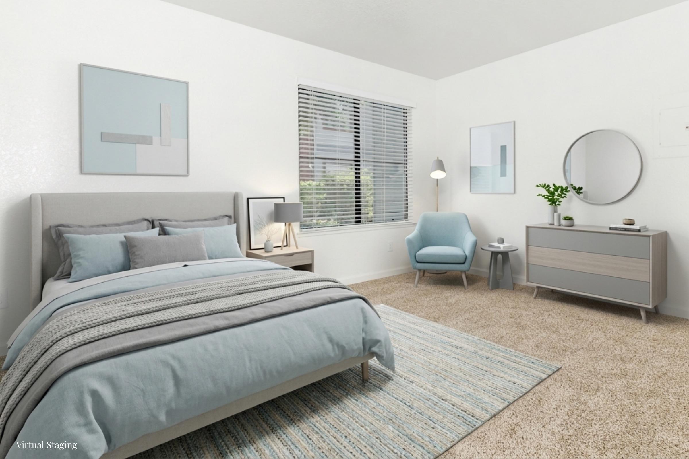 A modern, virtual-staged bedroom featuring a light gray bed with blue and gray bedding, a cozy armchair, a round mirror, and a stylish dresser. The walls are painted white, with minimalistic artwork and a window allowing natural light to illuminate the space. A soft area rug adds warmth to the carpeted floor.
