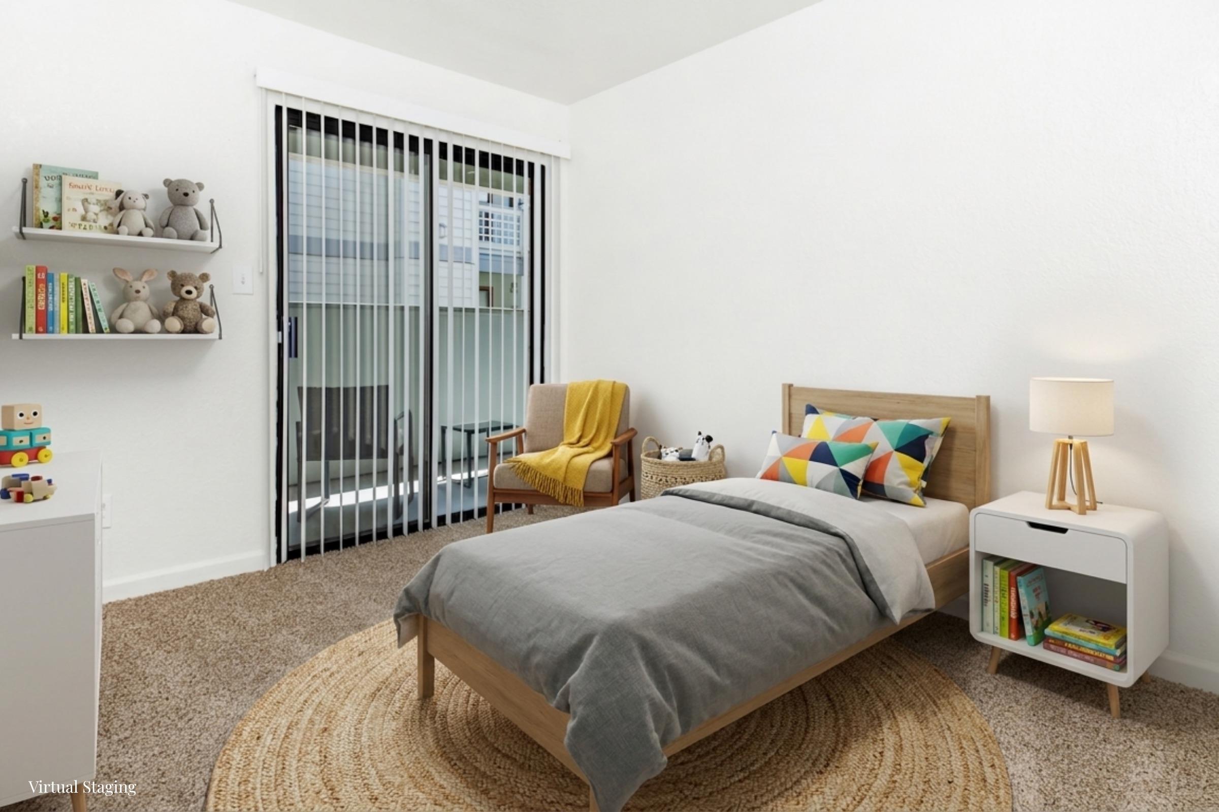A cozy bedroom featuring a twin bed with gray bedding and a colorful pillow. There’s a wooden nightstand with a lamp, a bookshelf filled with toys and books, and a comfortable armchair beside a sliding glass door. The room has light walls and a round area rug on a carpeted floor.