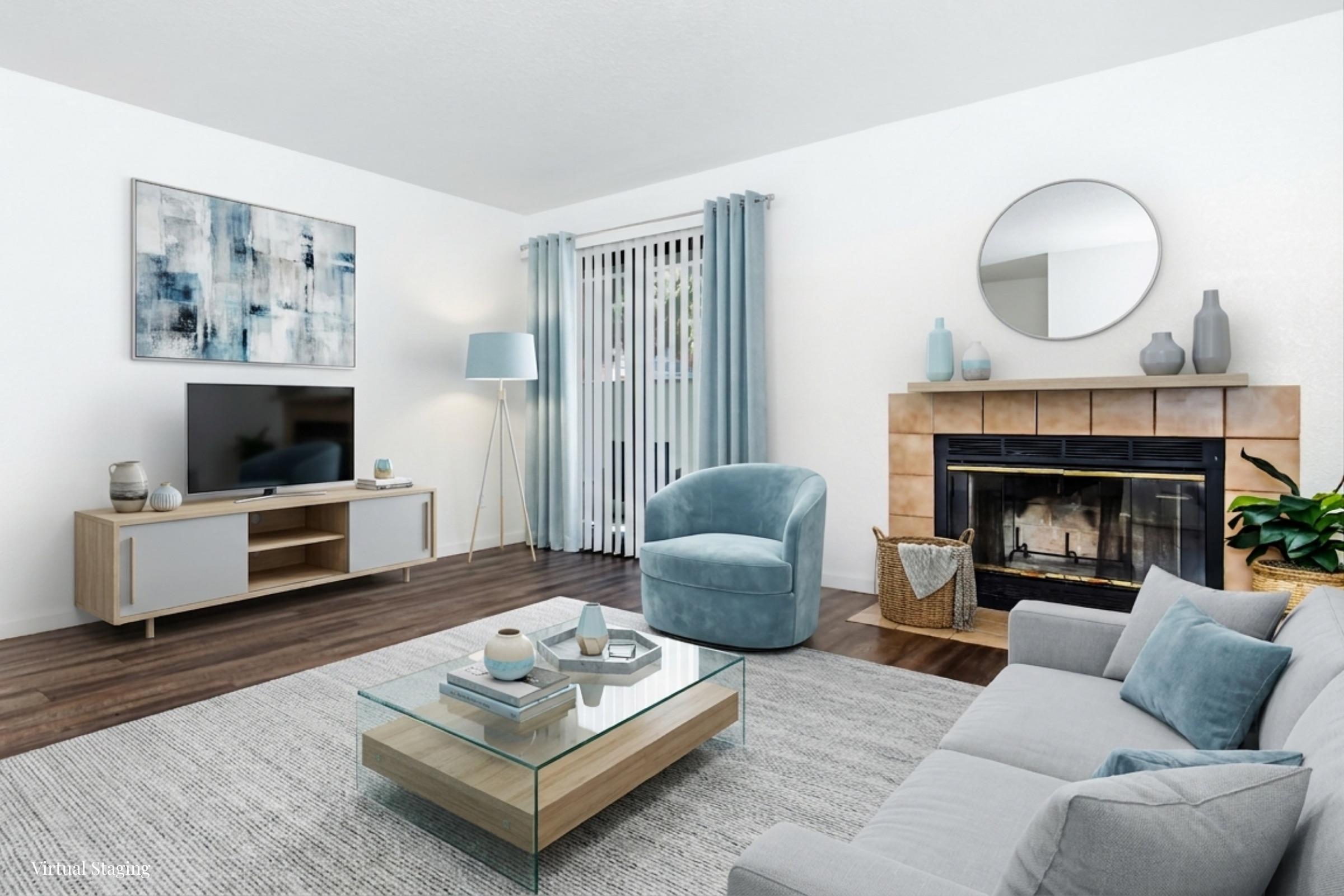A modern living room featuring a light gray sofa, a round blue armchair, and a glass coffee table on a textured rug. A wall-mounted TV sits on a wooden media console. There are blue curtains, a circular mirror above a stylish fireplace, and a potted plant in the corner, creating a cozy and inviting atmosphere.