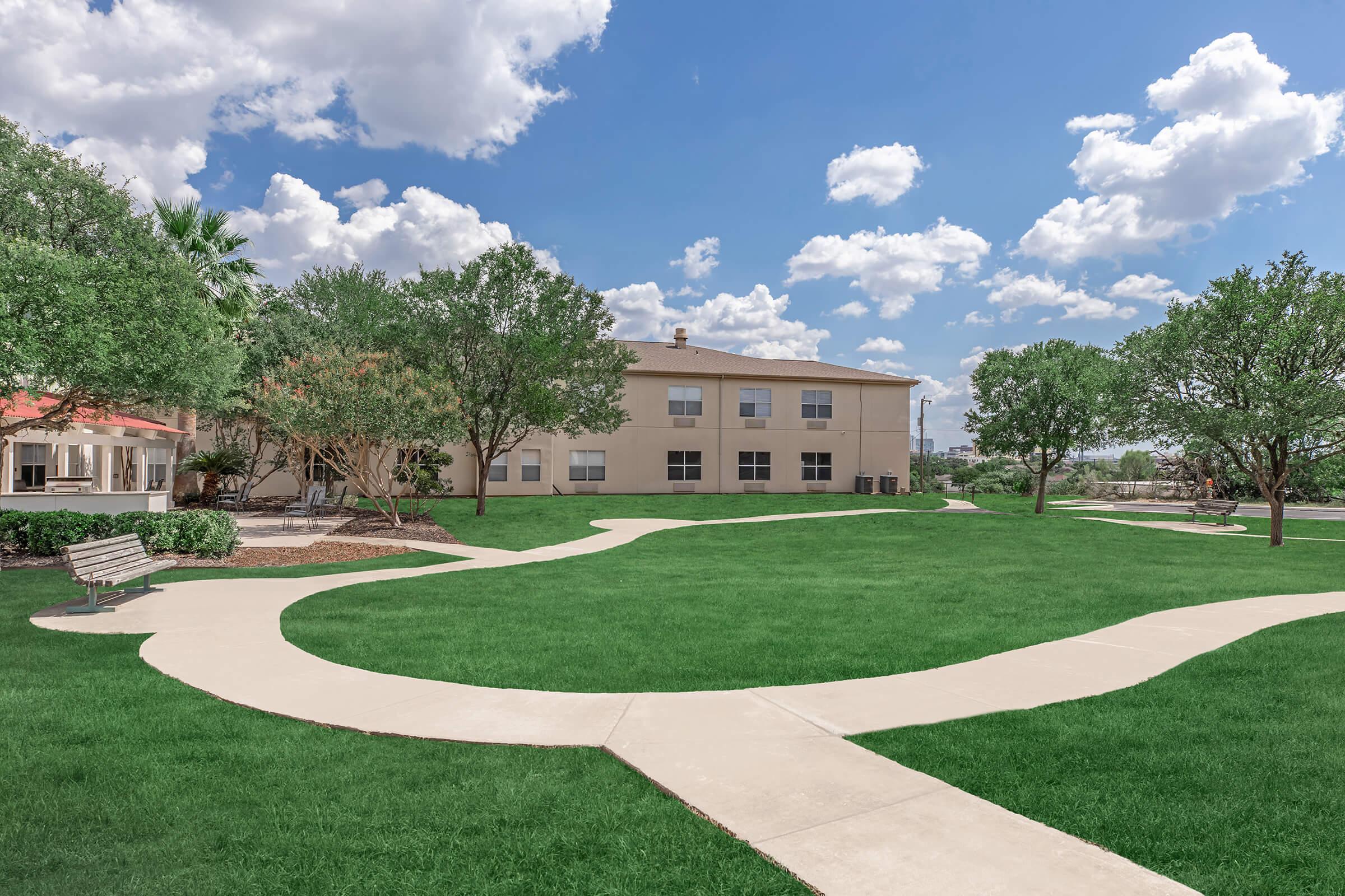 A landscaped outdoor area featuring a green lawn with winding concrete pathways. There are several trees and bushes providing shade, and a bench is positioned along the path. The background includes a beige building under a blue sky with fluffy white clouds.