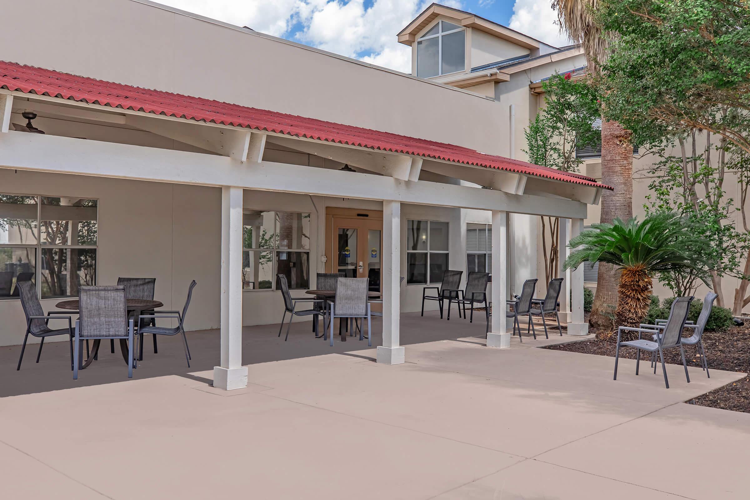 A shaded outdoor seating area with several metal tables and chairs under a white pergola. The background features a building with large windows and palm trees, with a clear blue sky and fluffy clouds above. The space is clean and inviting, suitable for relaxation or social gatherings.