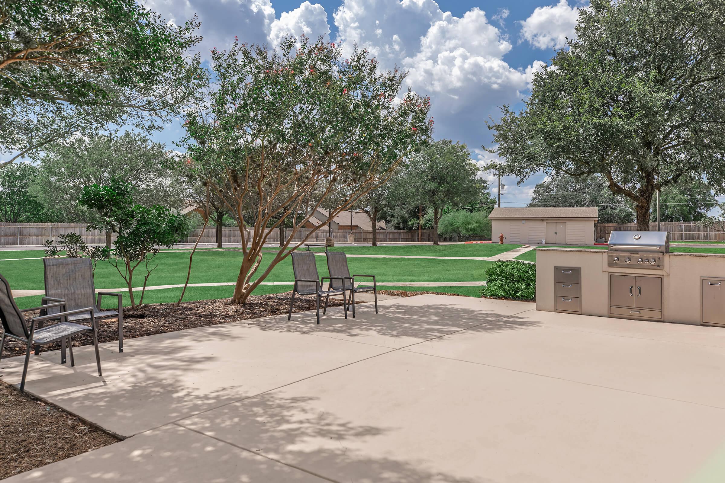 A spacious outdoor patio featuring several metal chairs, a built-in grill, and well-maintained landscaping. In the background, there are trees and a grassy area, with a clear blue sky populated by white clouds, providing a peaceful and inviting atmosphere for relaxation and outdoor cooking.