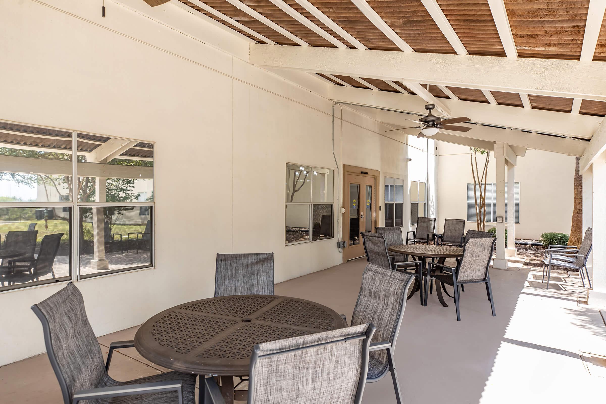 Outdoor patio area with multiple tables and chairs, shaded by a roof with wooden beams. Two ceiling fans and large windows reflect the surrounding greenery, creating a comfortable space for gatherings or relaxation. The flooring is smooth and light-colored, enhancing the inviting atmosphere.
