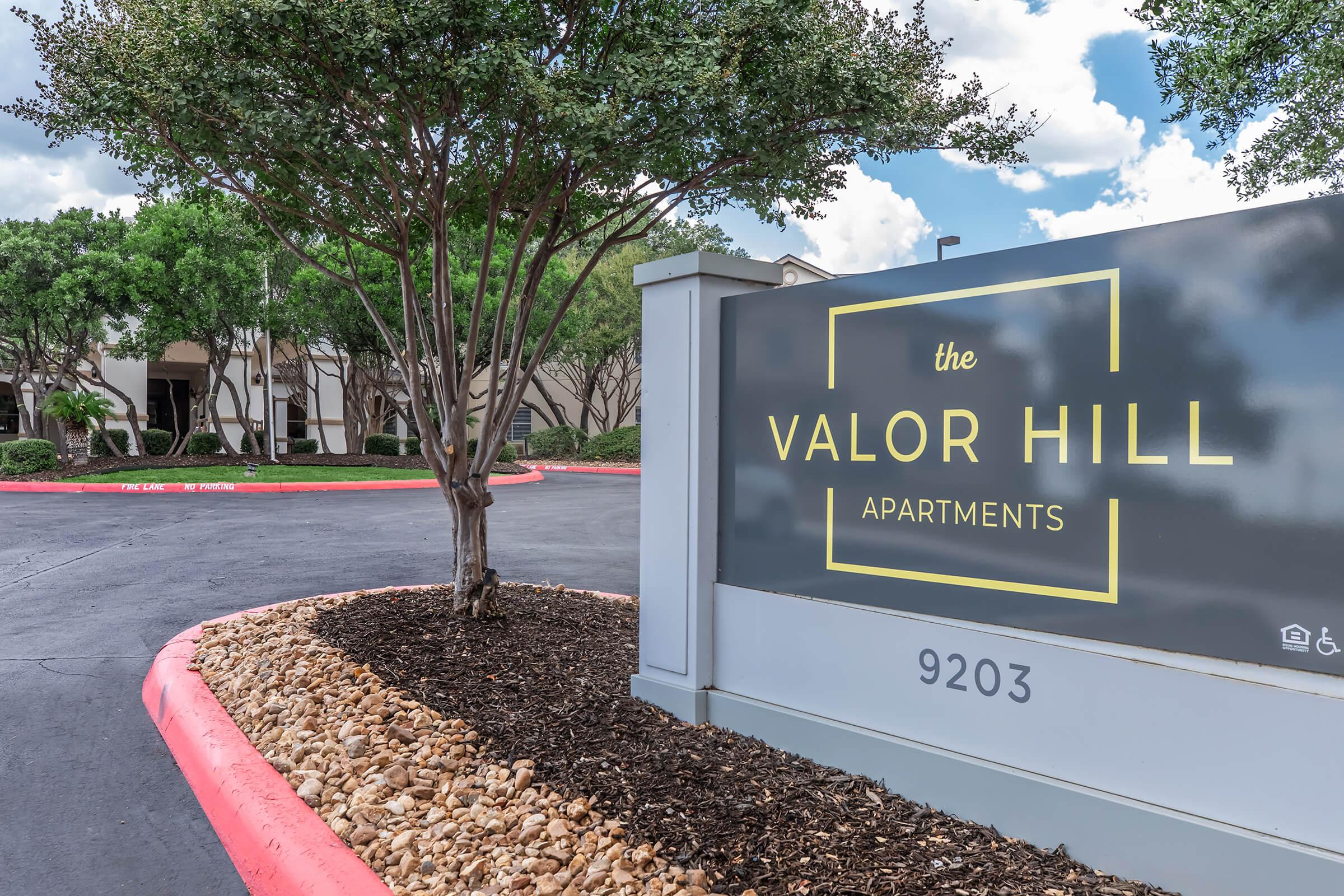 Sign for Valor Hill Apartments, featuring the name prominently displayed. The sign is positioned at the entrance of the complex, surrounded by a landscaped area with gravel and a tree. The background shows a building and a blue sky with fluffy clouds.