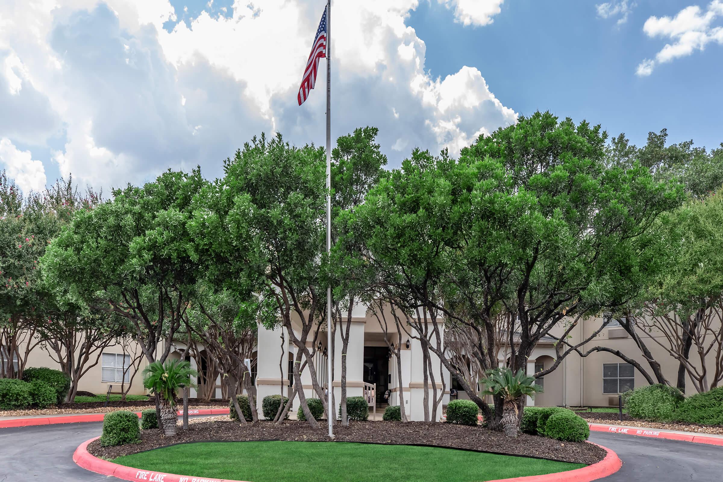 A landscaped entrance featuring several large green trees surrounding a flagpole with an American flag. The building behind has a light exterior and is partially obscured by the foliage. The sky above is partly cloudy, contributing to a bright, welcoming atmosphere.