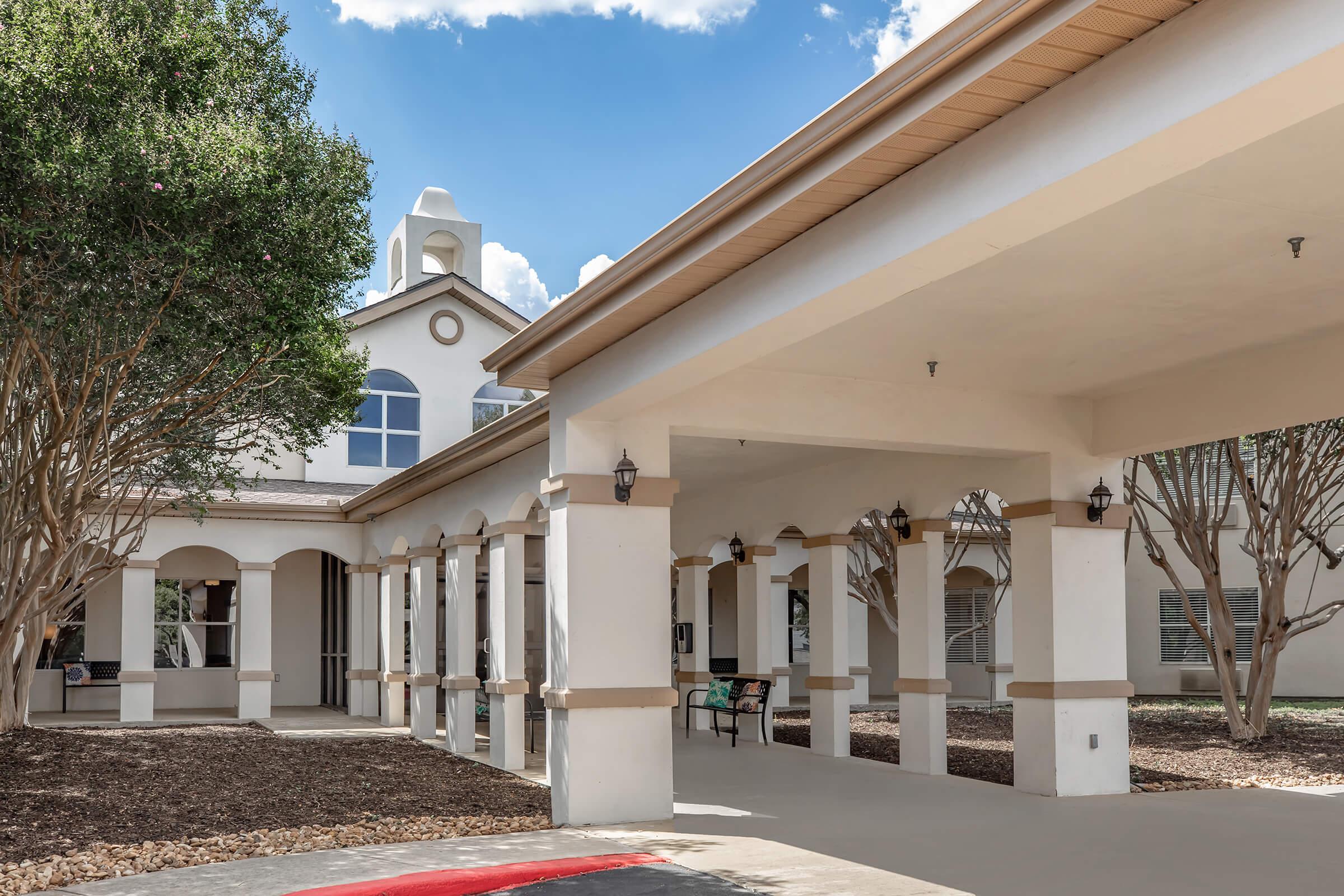 A modern building with a stucco exterior featuring arched columns, surrounded by trees and landscaping. The structure has a bell tower and large windows, with a covered walkway leading to the entrance. Blue sky and scattered clouds are visible above.