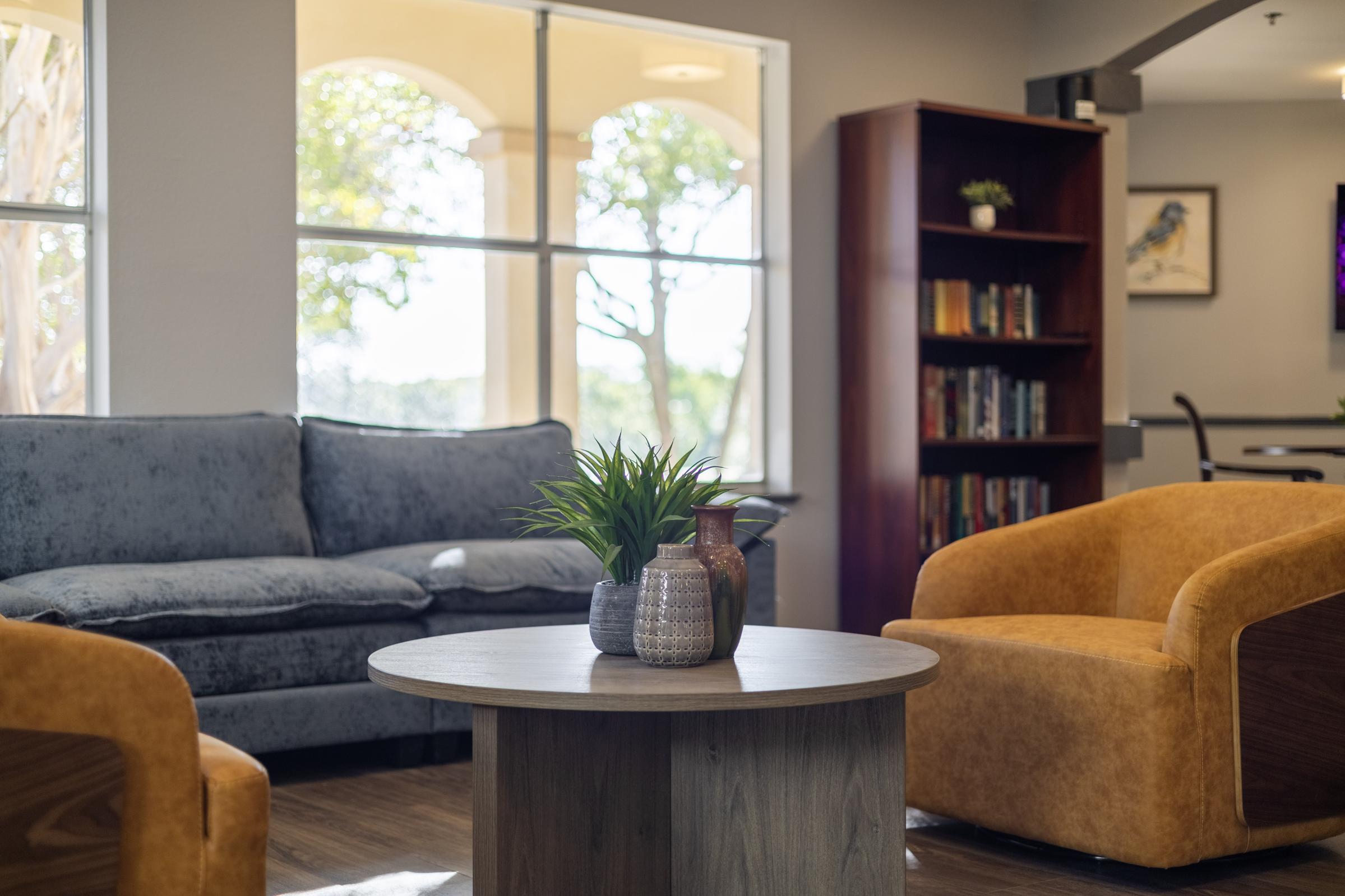 A cozy living room setting featuring a gray sofa, two orange accent chairs, and a round wooden coffee table. A small potted plant and a decorative vase sit on the table. In the background, a bookshelf filled with books and a large window with natural light and greenery outside.