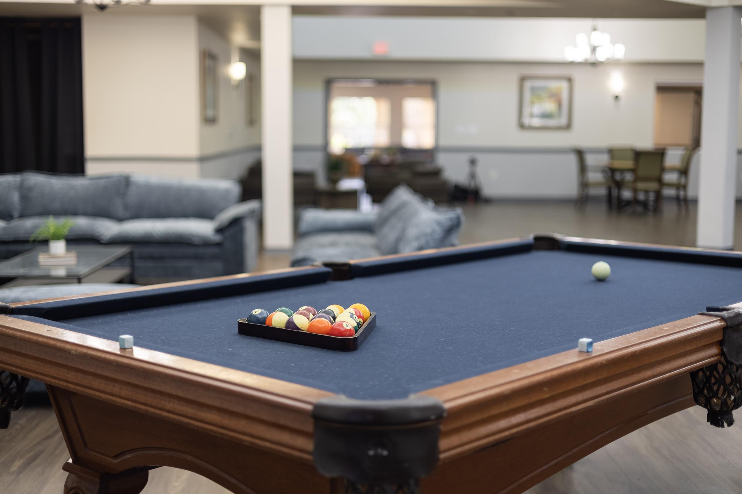 A close-up view of a pool table with a dark blue felt surface, featuring a tray of colorful billiard balls arranged on it. In the background, a lounge area with comfortable seating and tables is visible, suggesting a recreational space.