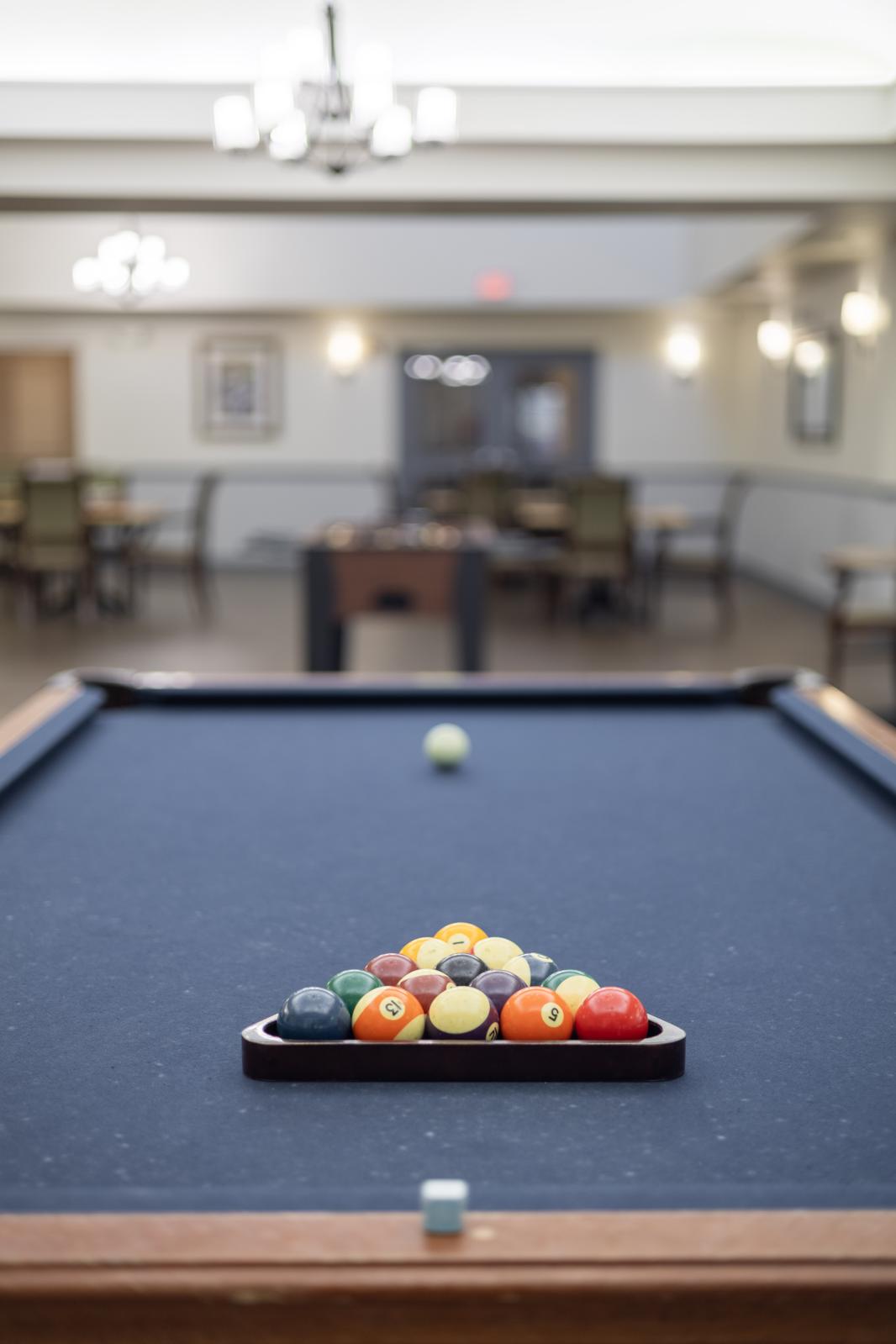 A pool table with a triangular rack of colored balls in the foreground. The table is set up in a spacious game room, with blurred tables and chairs in the background, illuminated by soft lighting.