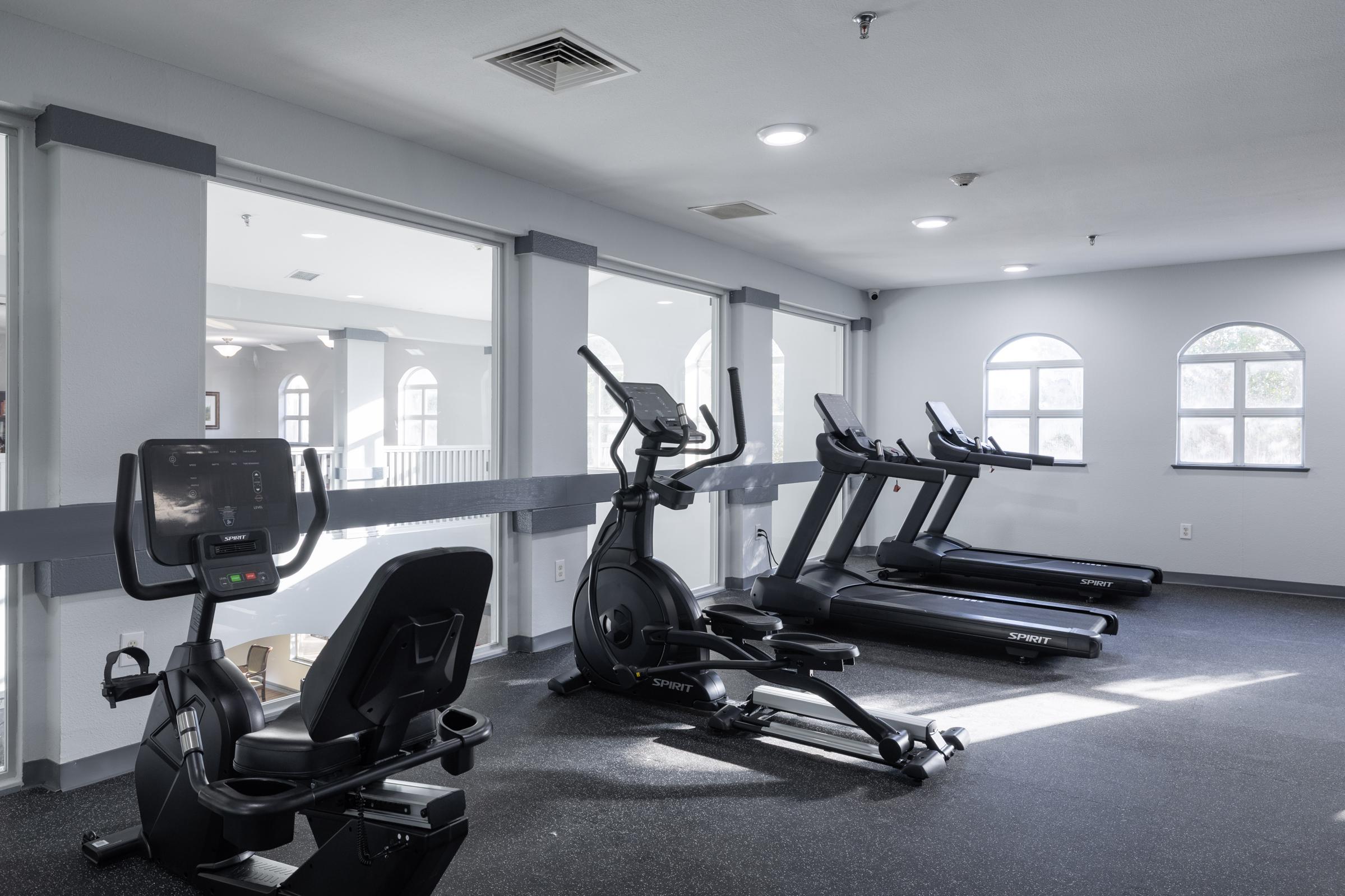 A bright and modern fitness room featuring a variety of exercise equipment, including an elliptical machine, a stationary bike, and multiple treadmills, all arranged on a black carpeted floor. Large windows provide natural light, and the walls are painted in soft neutral tones.