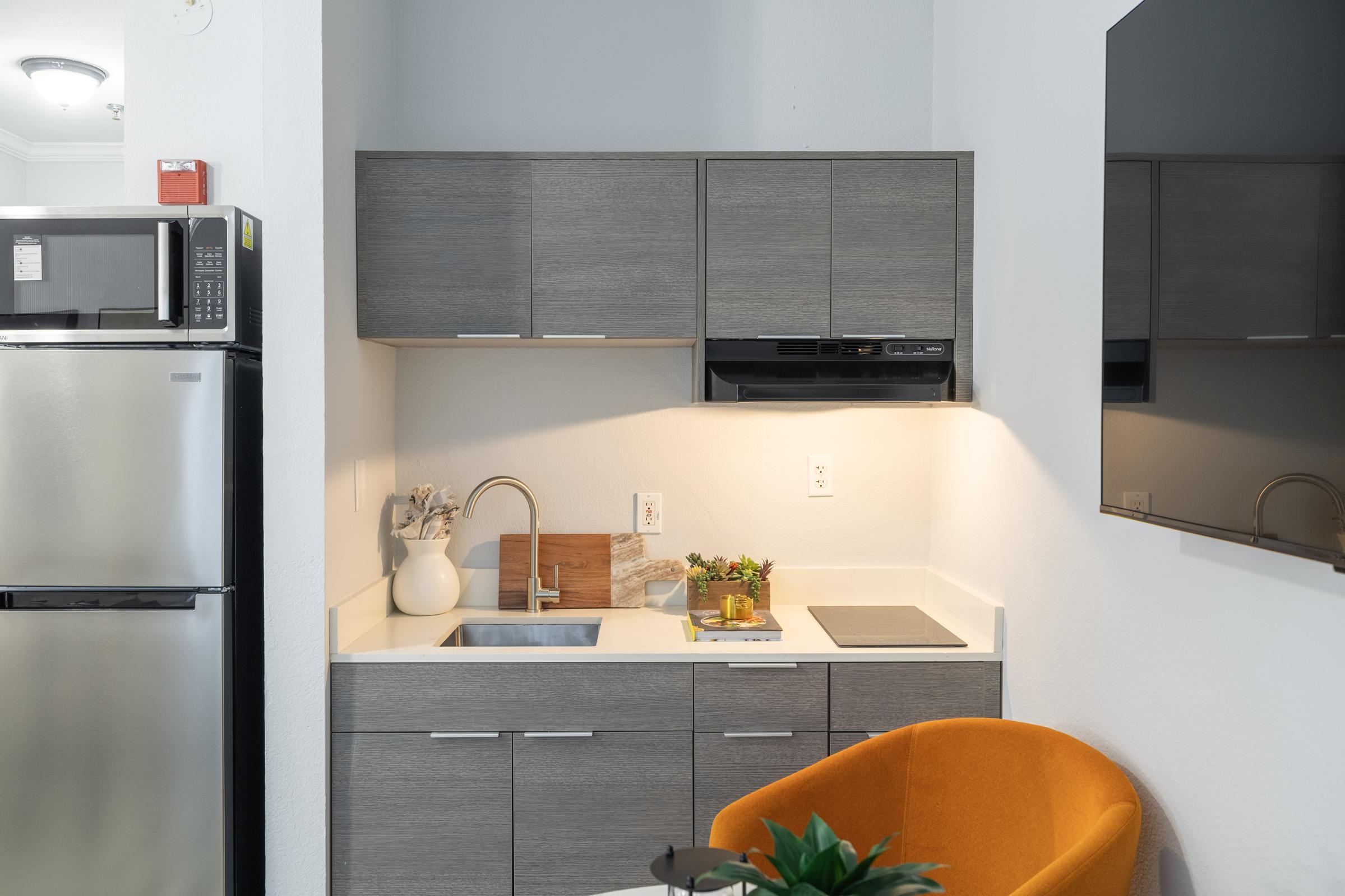 Modern kitchen space featuring a stainless steel refrigerator, gray cabinetry, a sink, a black microwave, and induction cooktop. There's a small wooden cutting board and some decorative items on the counter, along with a cozy orange chair and a small plant, creating a stylish and functional atmosphere.