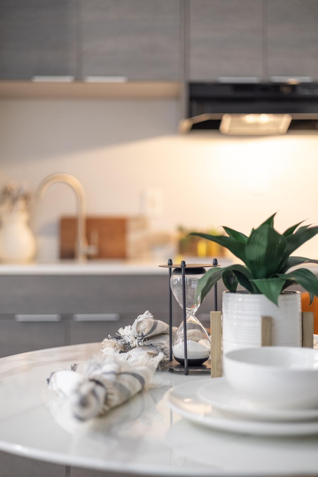 A stylish kitchen scene featuring a white dining table set with a simple plate and decorative items. An hourglass and a small potted plant add charm. The background shows a modern kitchen with gray cabinetry and warm lighting, creating a welcoming atmosphere.