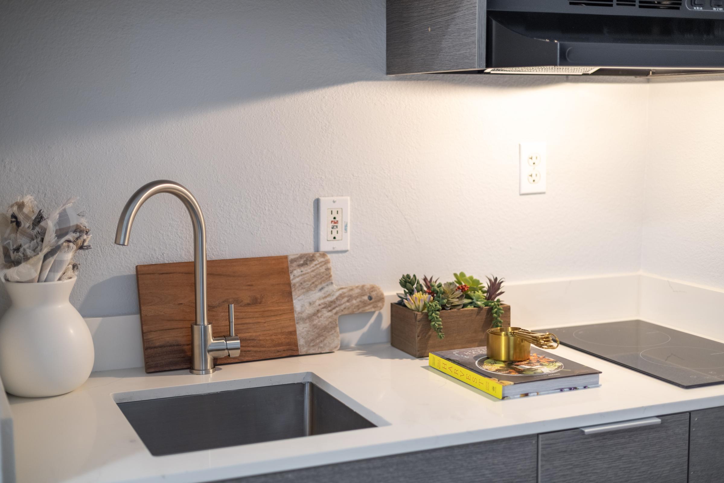 A modern kitchen countertop featuring a sink with a sleek faucet, a wooden cutting board, a decorative planter with succulents, a vase with kitchen utensils, and a stack of books. The space is well-lit with warm overhead lighting and has a minimalist design.