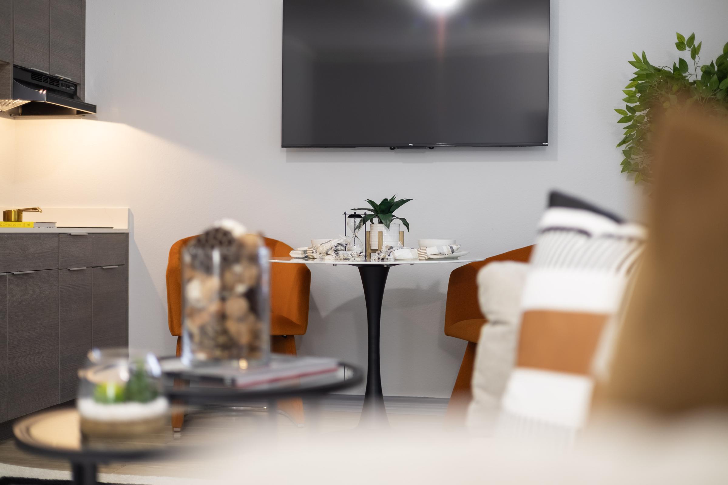 A cozy living space featuring a stylish dining table set with white dishware and a plant centerpiece. Two orange chairs are positioned around the table, while in the foreground, a round coffee table holds a glass jar filled with decorative items and a small plant. A flat-screen TV is mounted on the wall in the background.