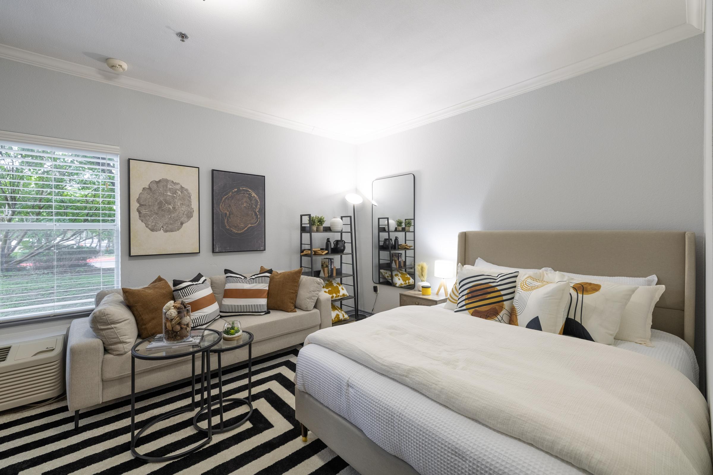 A cozy bedroom featuring a bed with decorative pillows, a stylish beige headboard, and a small seating area with a sofa. The room has a striped black-and-white rug, modern artwork on the walls, a round coffee table with decor, and a standing lamp, all illuminated by natural light from a window.