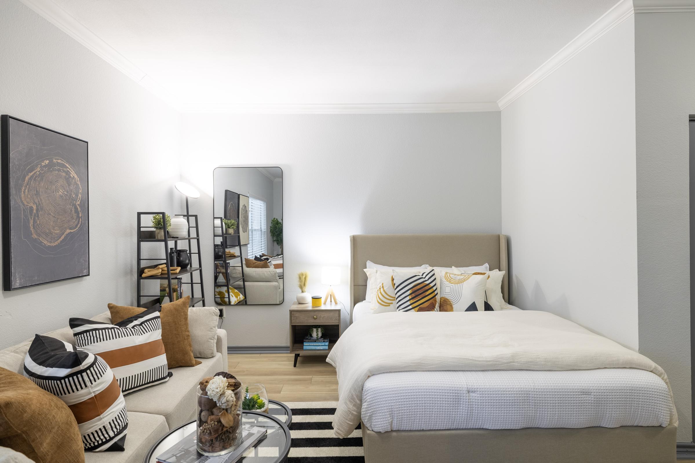A modern bedroom featuring a cozy bed with white linens, decorative pillows, and a light-colored headboard. There’s a stylish mirror and a nightstand beside the bed. A small sofa with decorative pillows sits next to a glass coffee table adorned with a decorative centerpiece. The walls are light gray, creating a fresh ambiance.