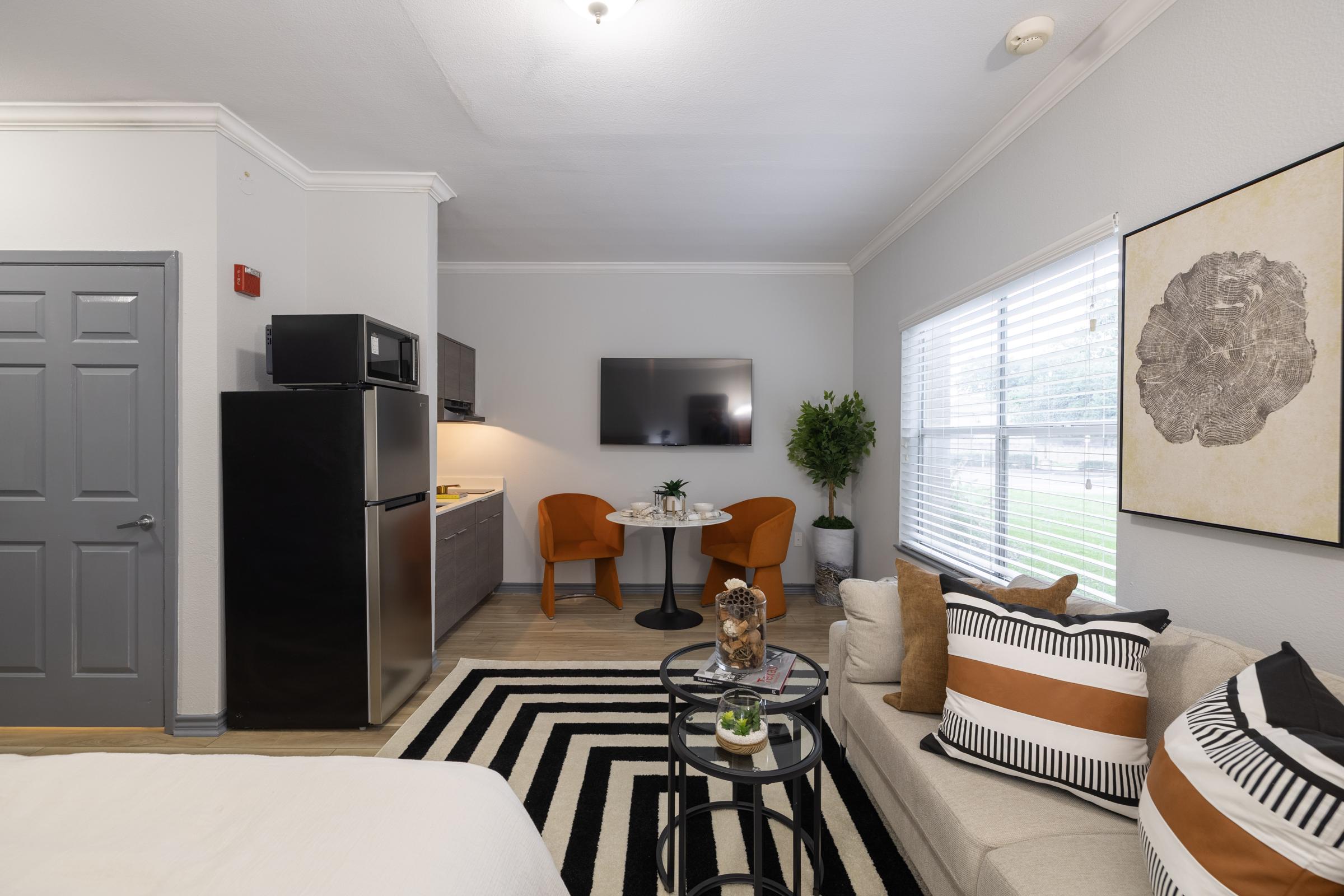 A modern living space featuring a small dining area with two orange chairs and a round table, a kitchenette with a black fridge, and a cozy sofa adorned with patterned pillows. The room has light-colored walls, large windows with blinds, and a decorative plant enhancing the ambiance.