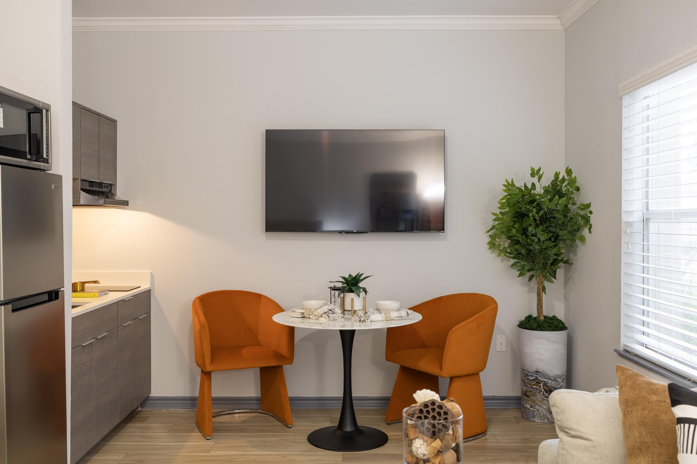 A cozy dining area featuring two orange chairs around a circular black table, set with decorative items. A modern kitchen with gray cabinetry is visible on one side, and a wall-mounted TV is on the opposite side. A green potted plant adds a touch of nature, while natural light comes through a window.