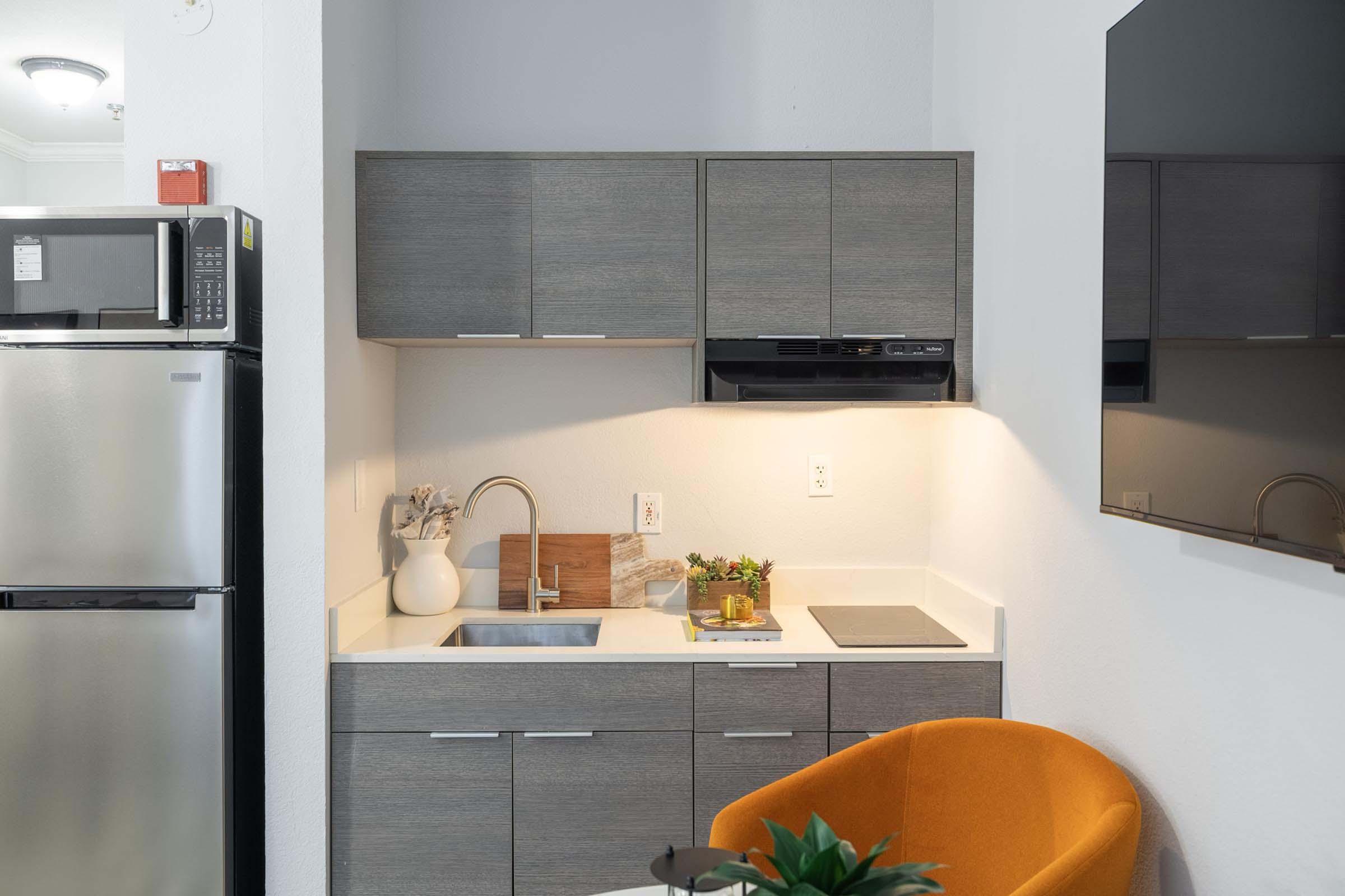 A modern kitchen area featuring gray cabinets, a stainless steel refrigerator, and a small sink. There's a wooden cutting board and decorative plant on the countertop. An orange chair is positioned nearby, and a wall-mounted TV is visible. The overall design is sleek and minimalistic.
