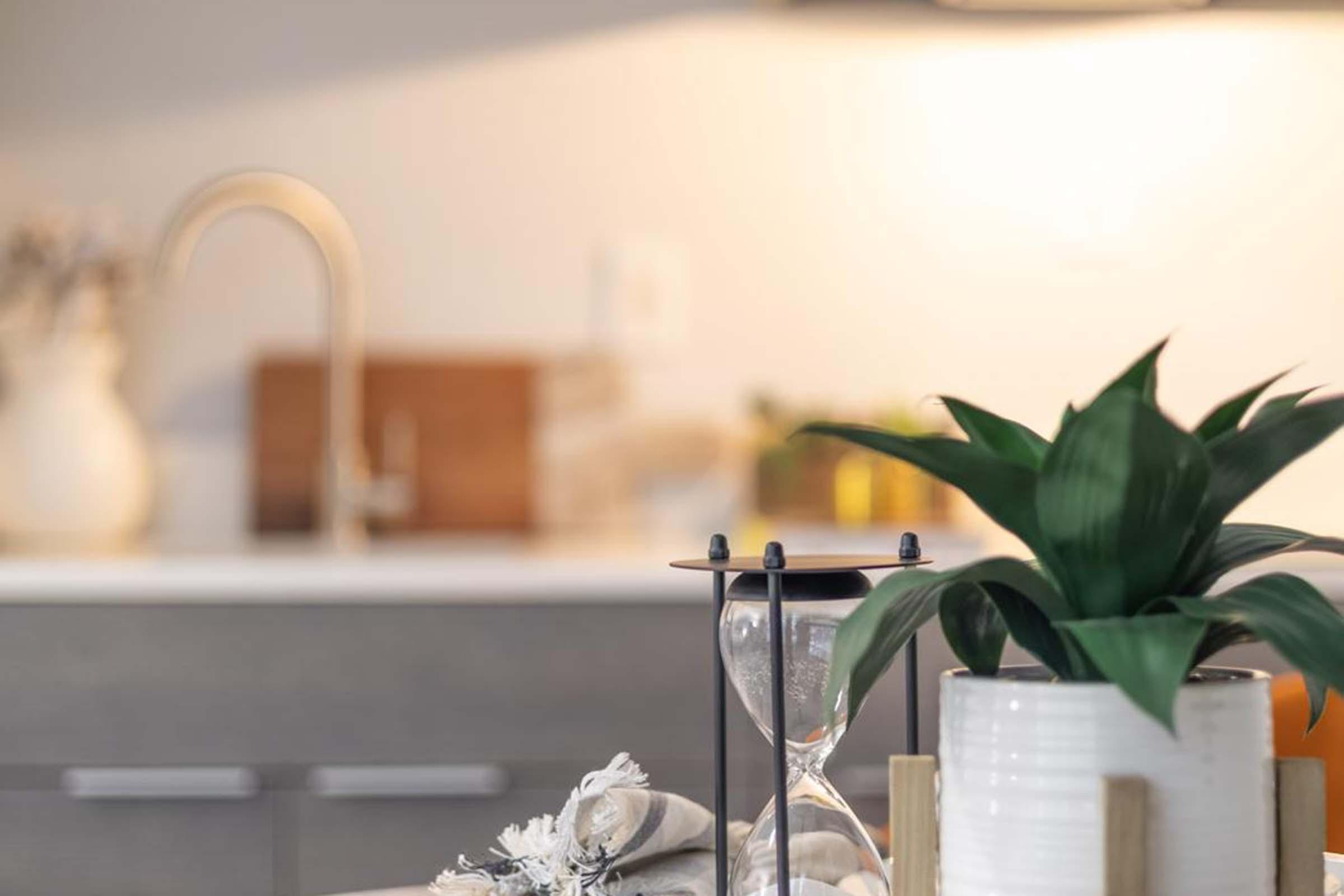 A close-up view of a kitchen countertop featuring a decorative hourglass, a potted plant, and a minimalistic candle holder. The background is softly blurred, highlighting the modern kitchen design with wooden accents and a stainless steel faucet.