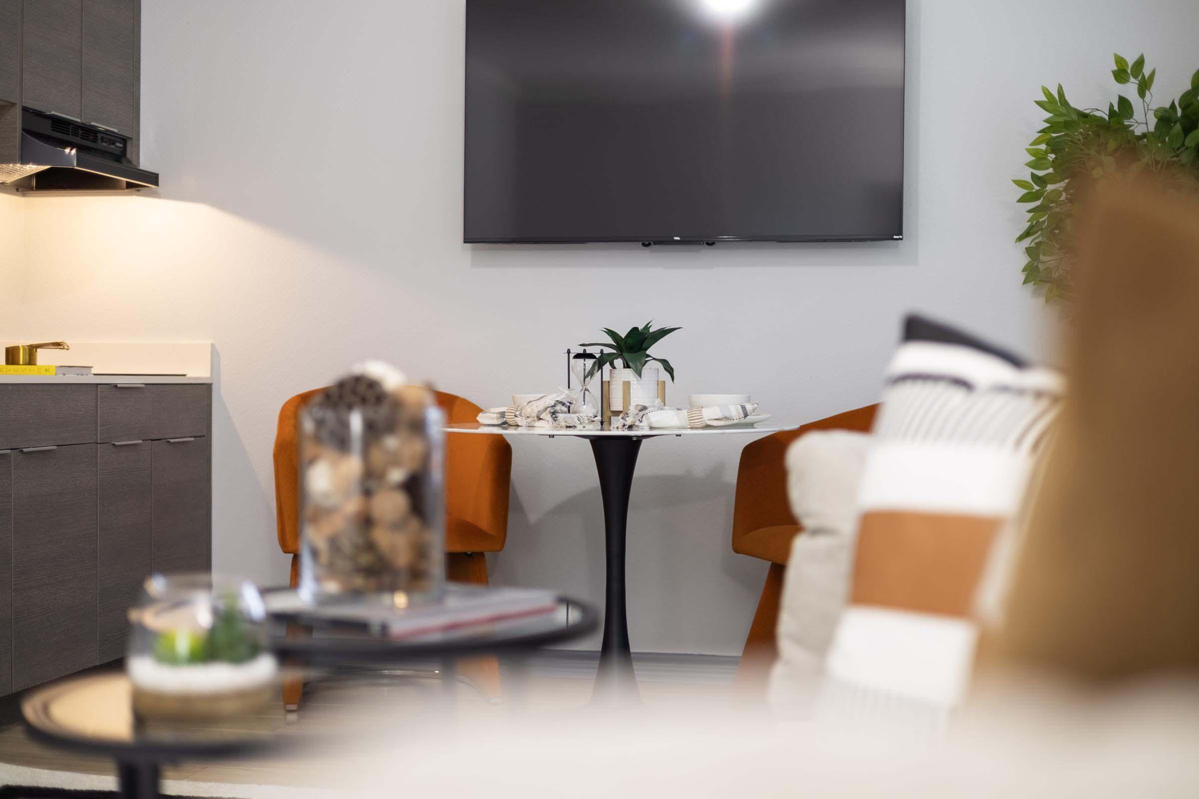A cozy living space featuring a modern design, including a wall-mounted TV, a small round table set with dishes and a plant, and two orange chairs. A coffee table in the foreground holds decorative items, while a soft, neutral-colored sofa adds warmth to the room. Natural light brightens the inviting atmosphere.