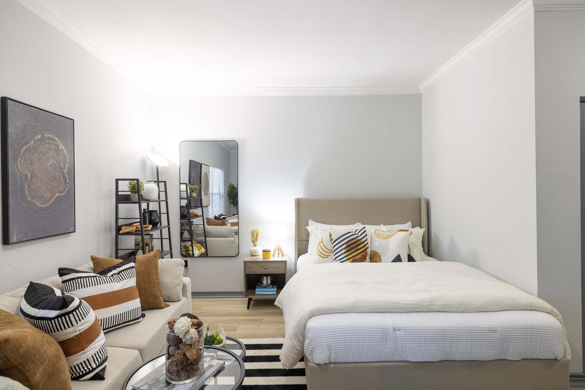 A cozy bedroom featuring a beige bed with decorative pillows, a stylish sofa adorned with patterned cushions, a round coffee table with decorative items, and a full-length mirror. The walls are light gray, and there is a small bookshelf with plants and decorative objects, creating a modern and inviting atmosphere.