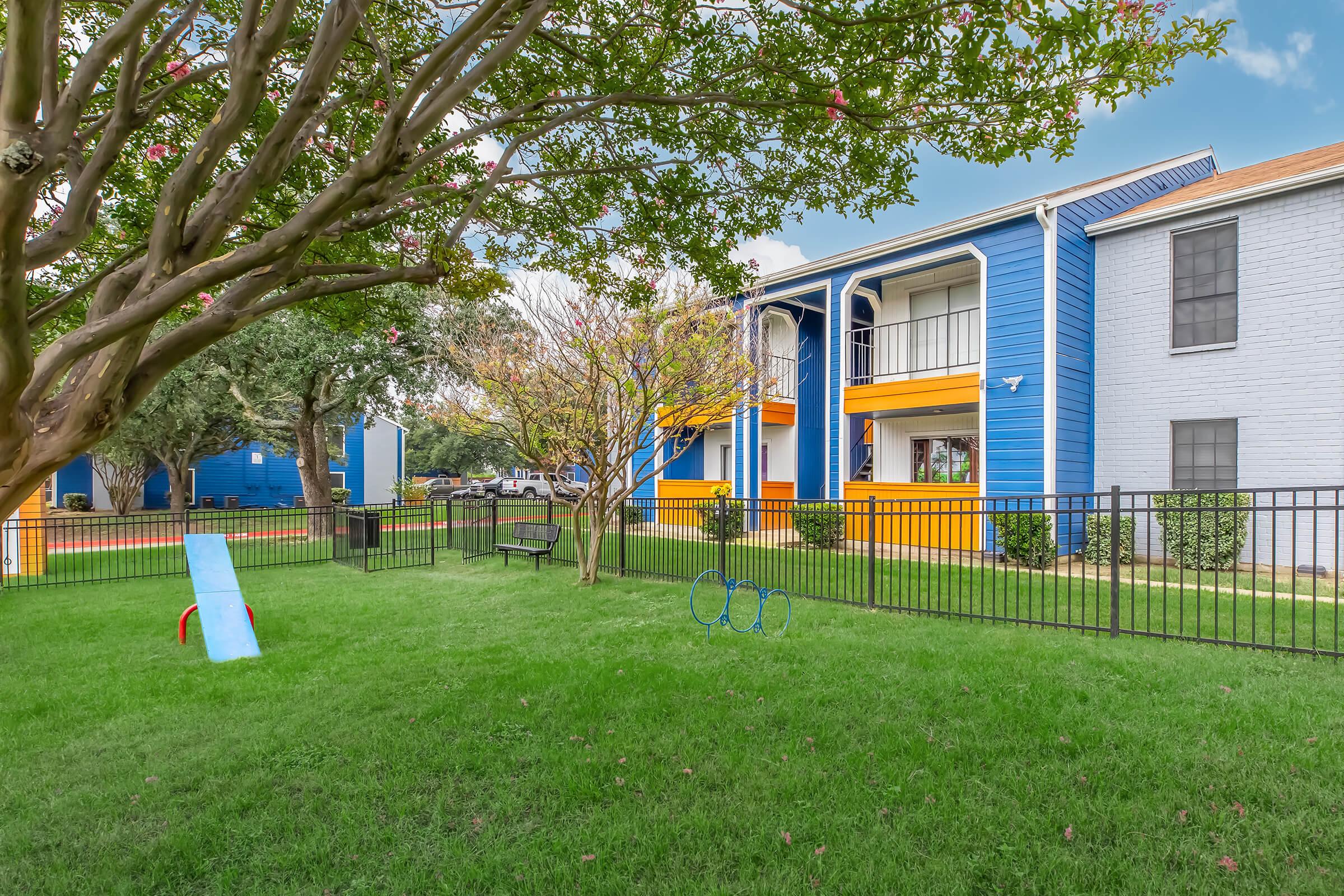 Colorful apartment complex with blue and orange buildings, surrounded by a well-maintained green lawn. There are trees, a small playground with a slide and climbing equipment, and a black fence enclosing the area. The sky is clear, contributing to a pleasant outdoor atmosphere.