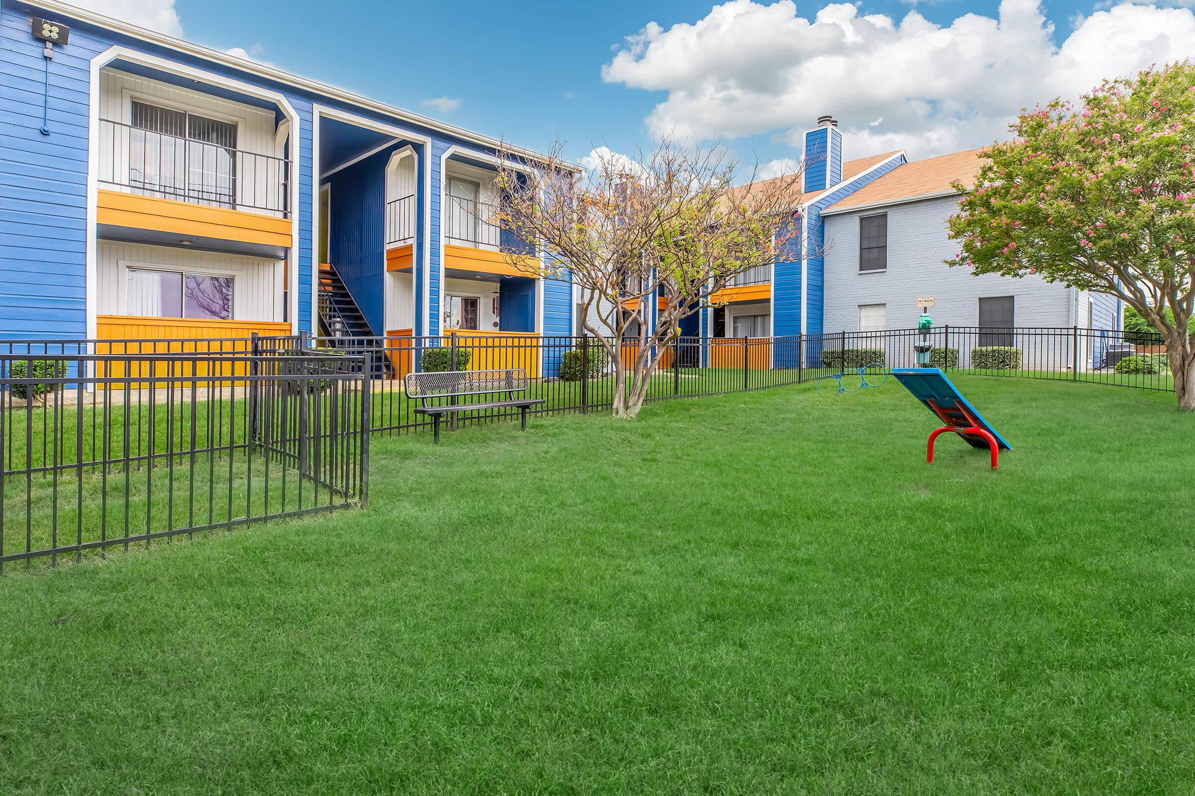 Colorful two-story apartment buildings surrounded by green grass and trees. A playground with a slide is visible in the foreground, along with a bench and a fence enclosing the area. The sky is partly cloudy, adding a bright atmosphere to the scene.