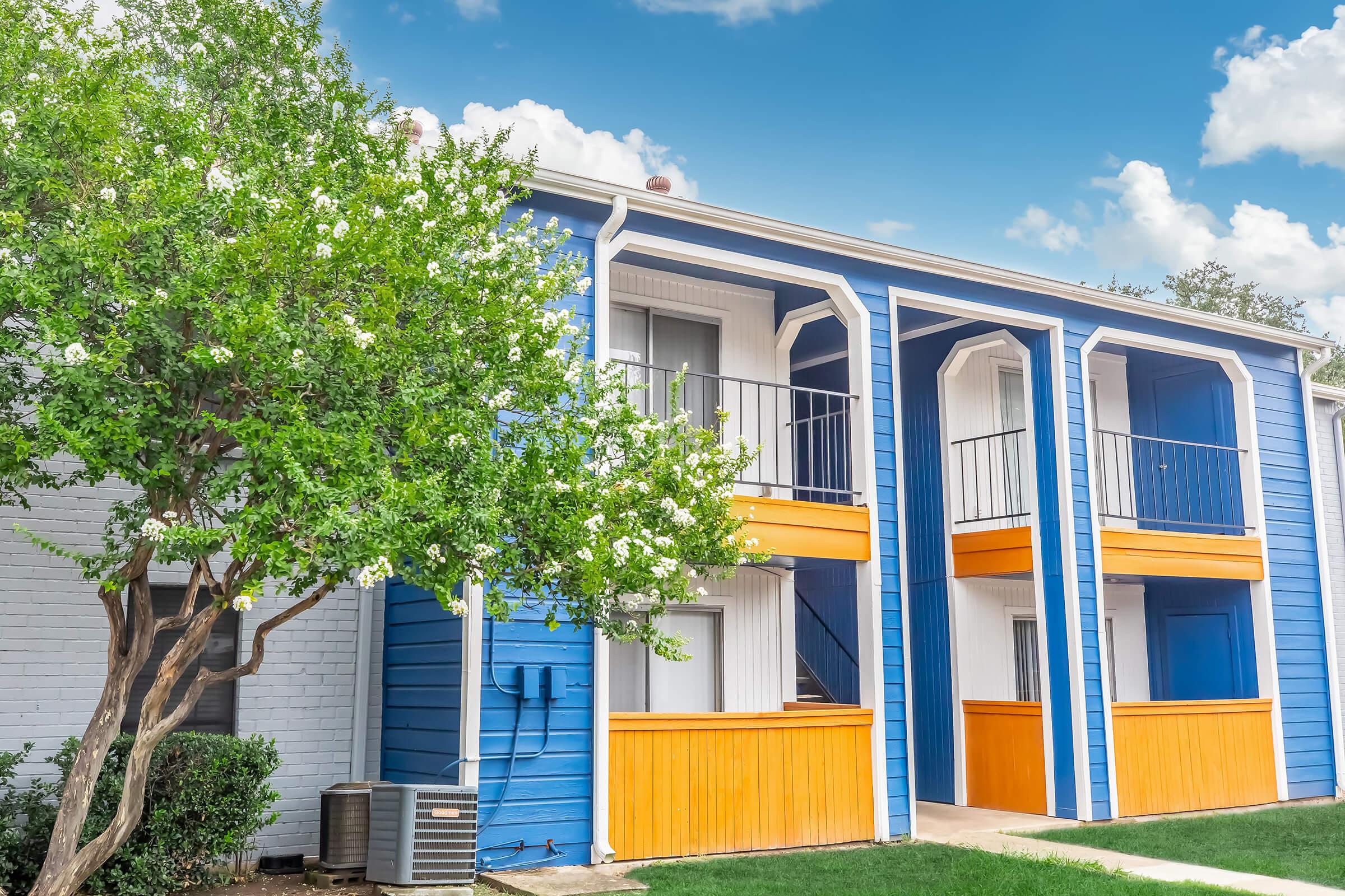 Two-story apartment building with blue and orange accents. The structure features balconies with railings on each level. A small tree with white flowers is in front of the building, and there is a grassy area in the foreground. Bright blue sky with some clouds in the background.