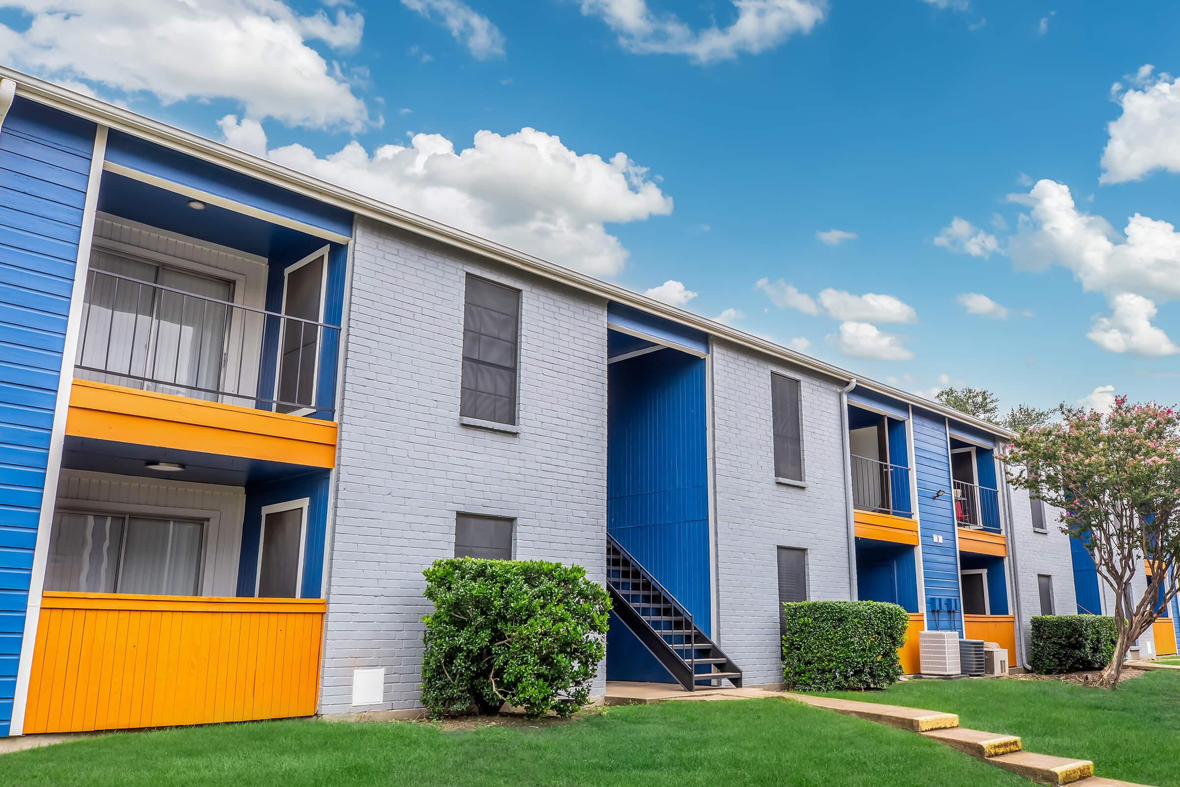 Apartment building with a modern design featuring blue and orange accents. The two-story structure has multiple balconies and large windows, surrounded by green lawns and neatly trimmed hedges. A staircase leads to the upper floor, against a backdrop of a clear blue sky with fluffy white clouds.