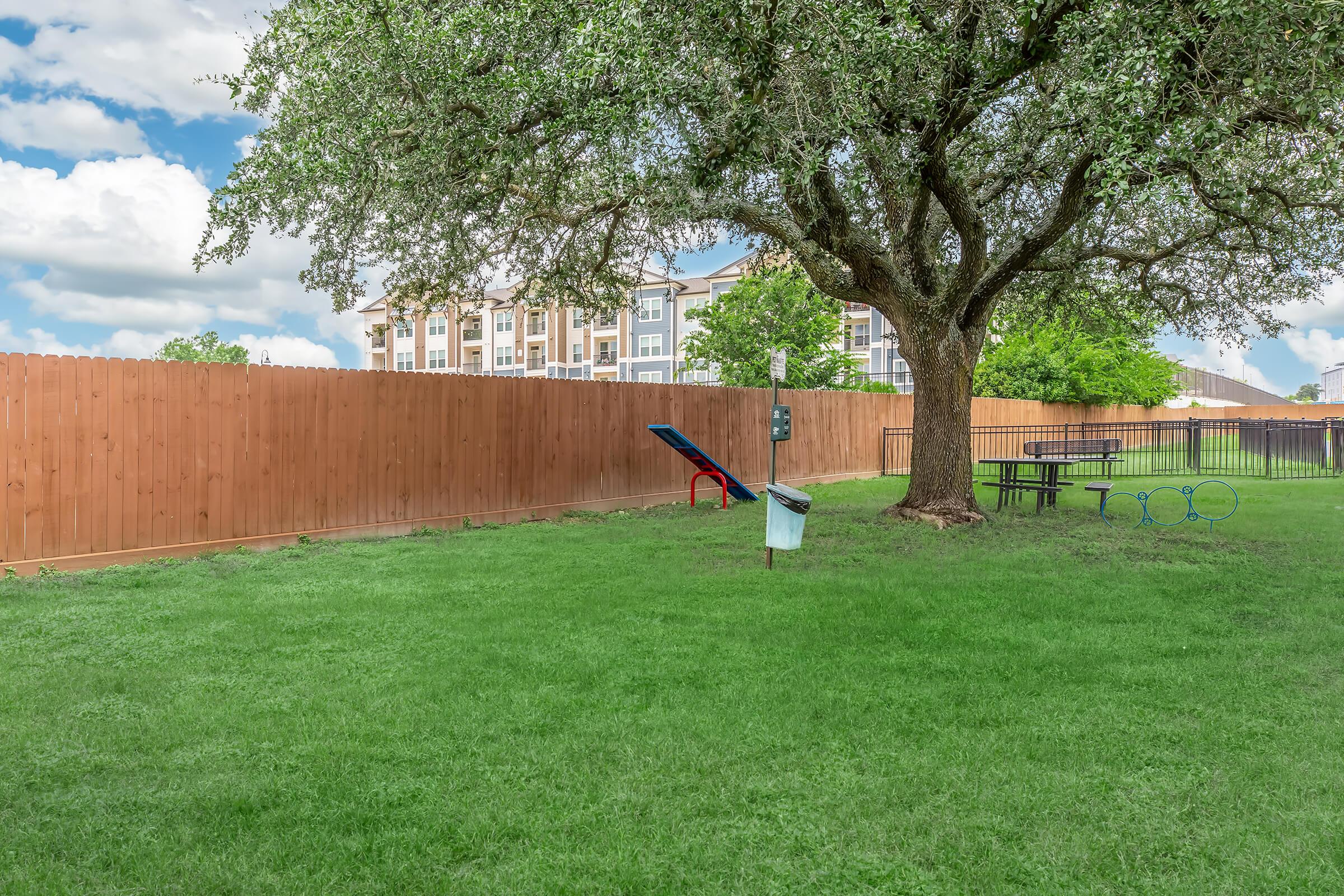 A grassy area with a large tree in the foreground, a wooden fence in the background, and an apartment building visible beyond the fence. There's a slide and a picnic table nearby, along with a trash bin and a pet waste station. The sky is partly cloudy, adding to the serene outdoor atmosphere.