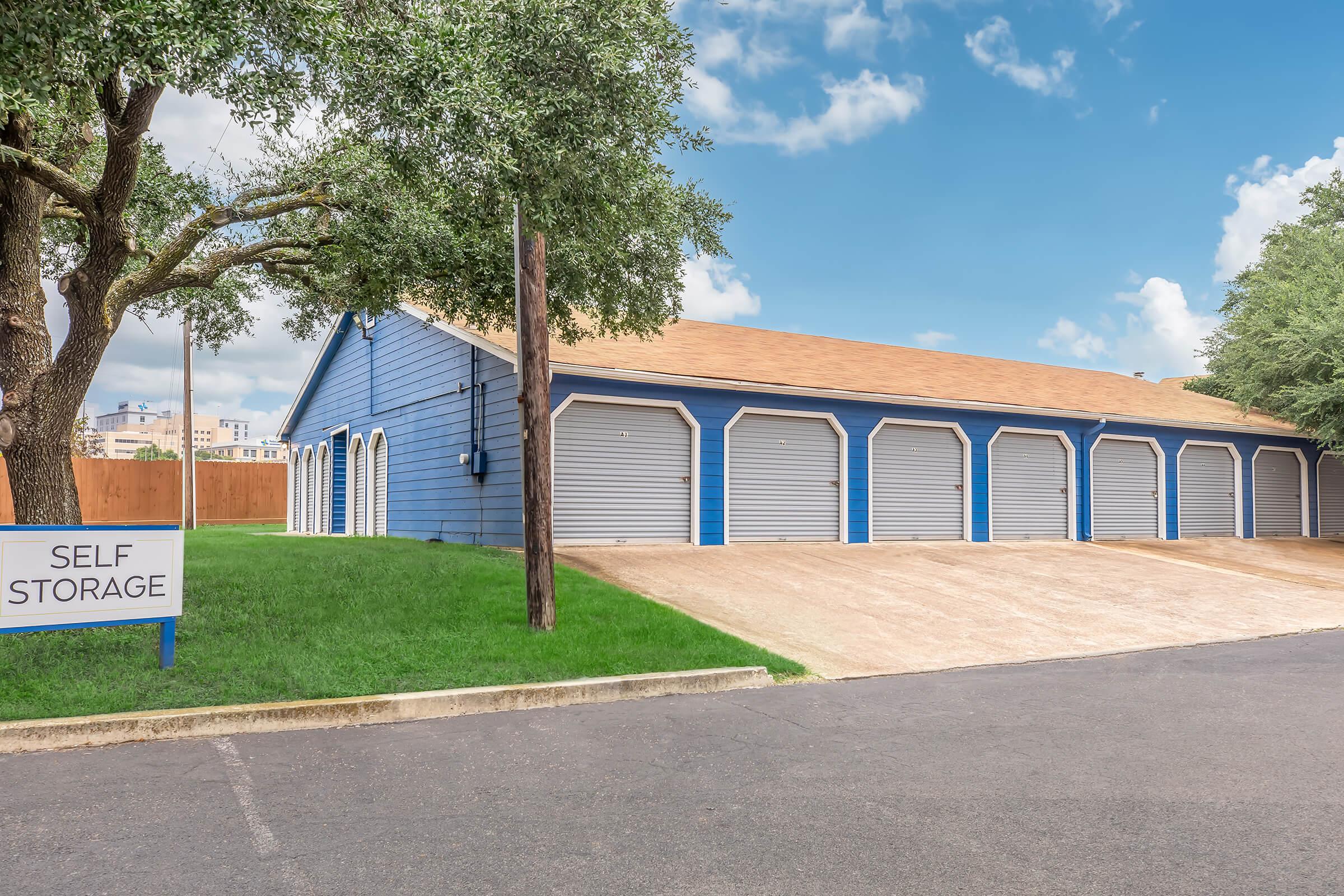 A blue self-storage building with multiple garage-style doors, set against a bright blue sky with a few clouds. There is green grass in front and a sign indicating "SELF STORAGE" next to the entrance. Nearby trees provide some shade, and the area is well-maintained.