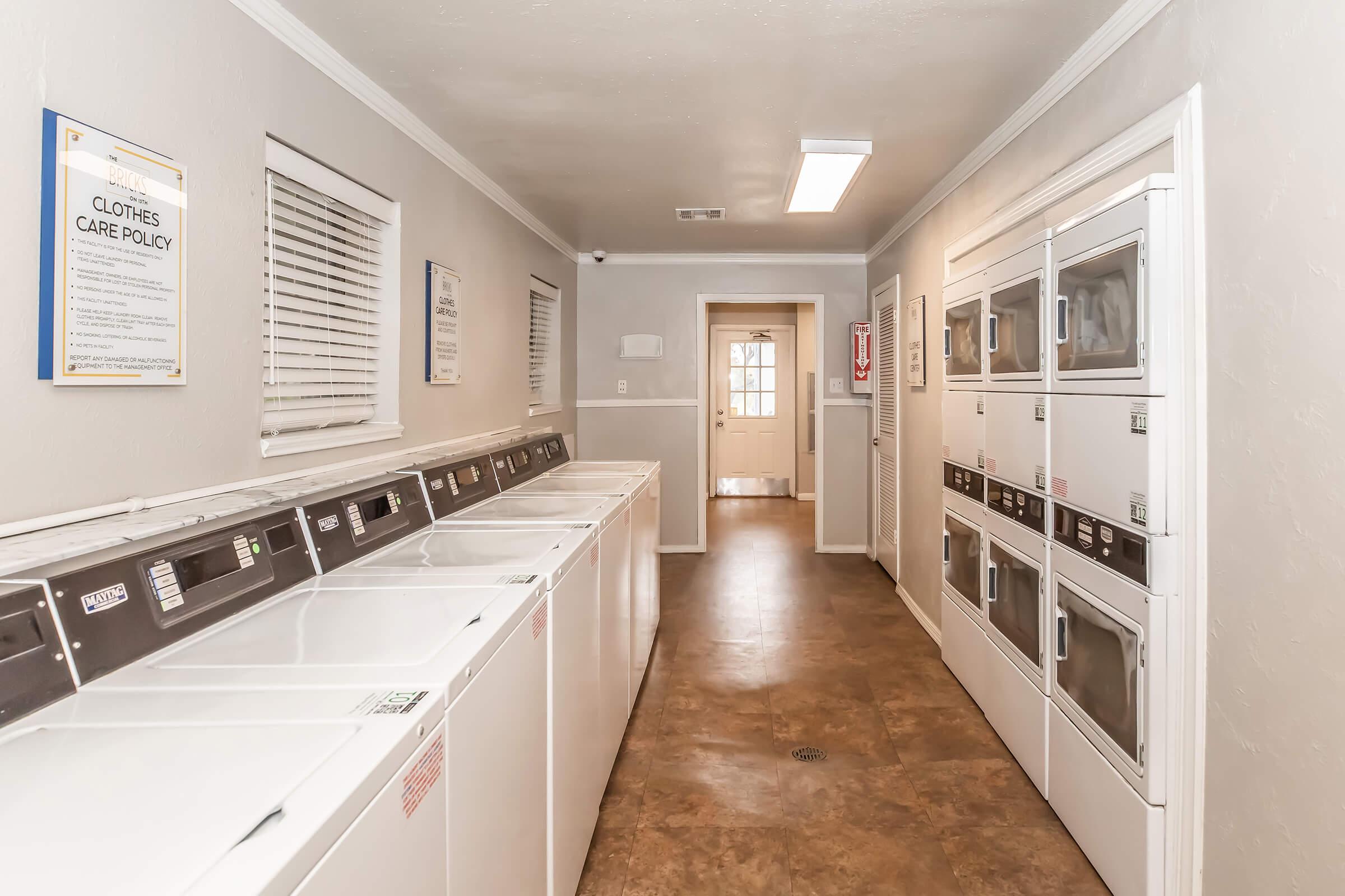 A clean and well-lit laundry room featuring multiple stacked washing machines and dryers along the walls. There are informational signs on the walls and a door at the far end leading to another area. The floors are polished and there are windows allowing natural light in.