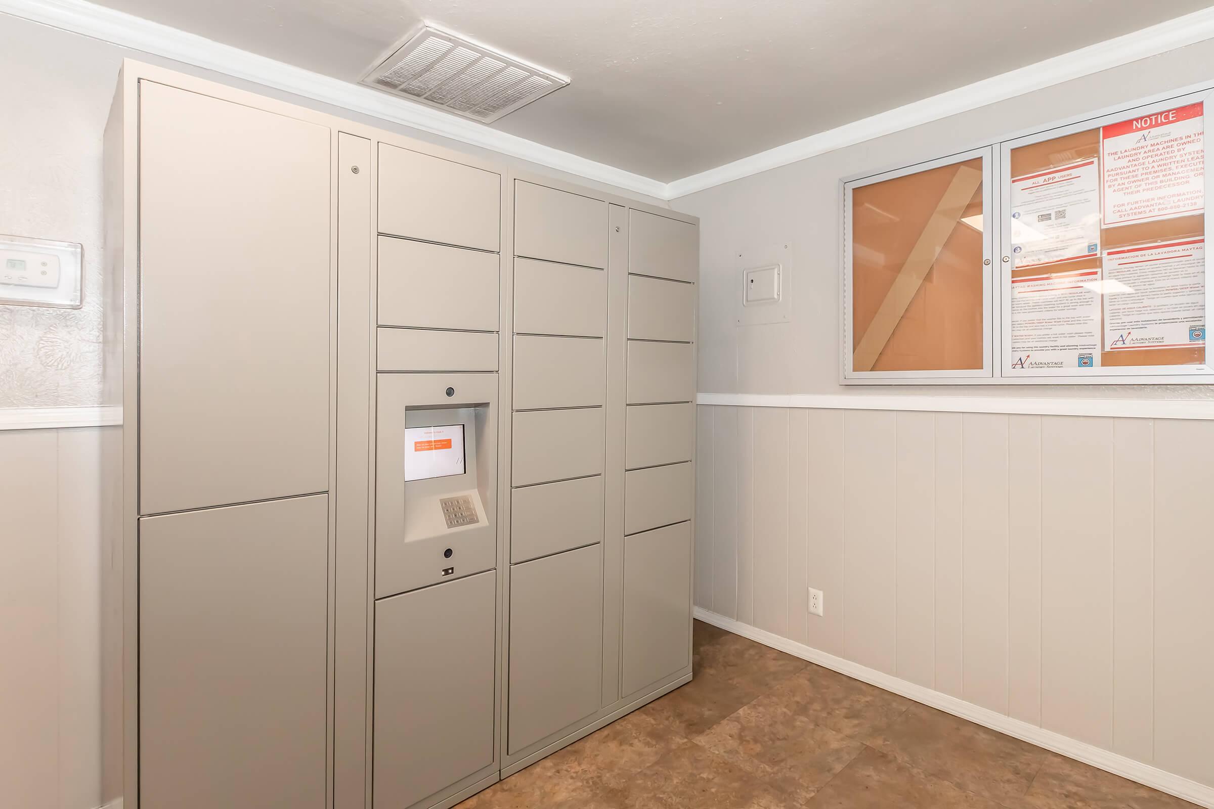 A secure package delivery locker system located in a well-lit room. The lockers are gray and arranged in a tall configuration, with a digital interface at the center for retrieving packages. A bulletin board with notices is mounted on the wall nearby, and the flooring features a tile pattern.