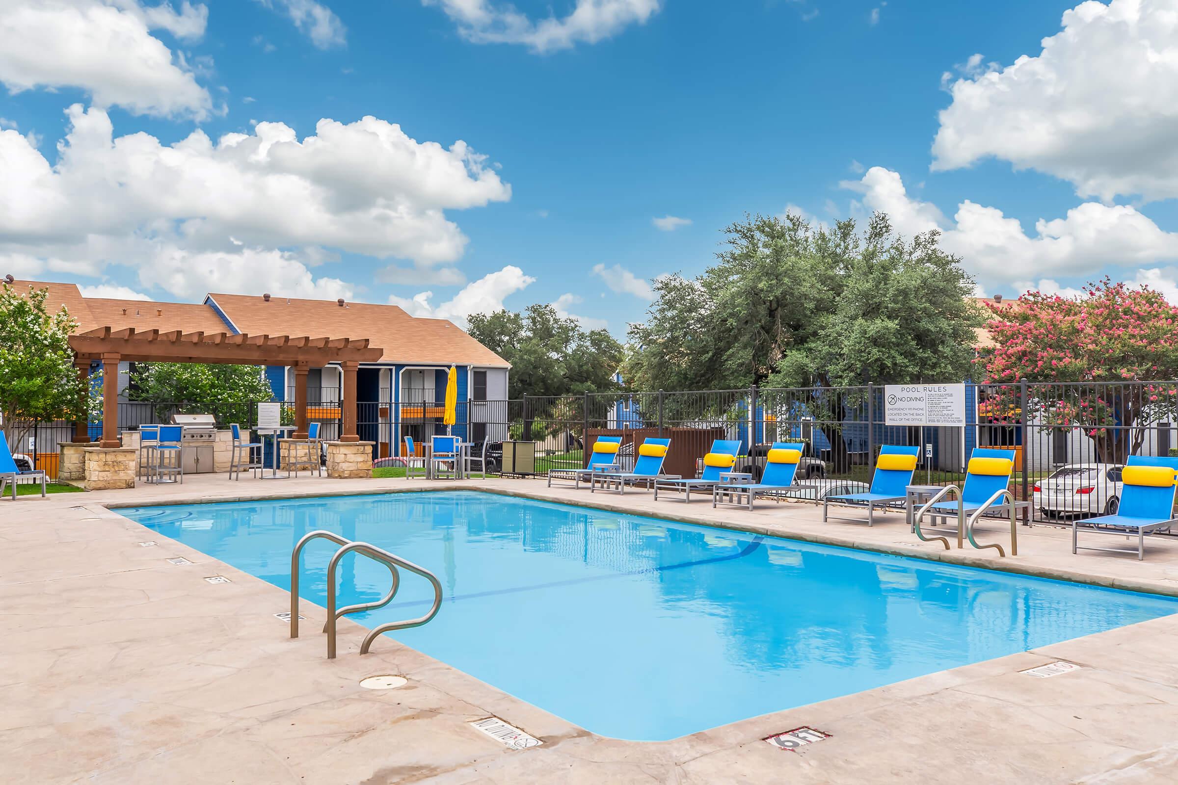 A bright outdoor swimming pool surrounded by lounge chairs under a sunny sky with fluffy clouds. In the background, there are trees and buildings, creating a relaxing atmosphere for leisure and enjoyment.