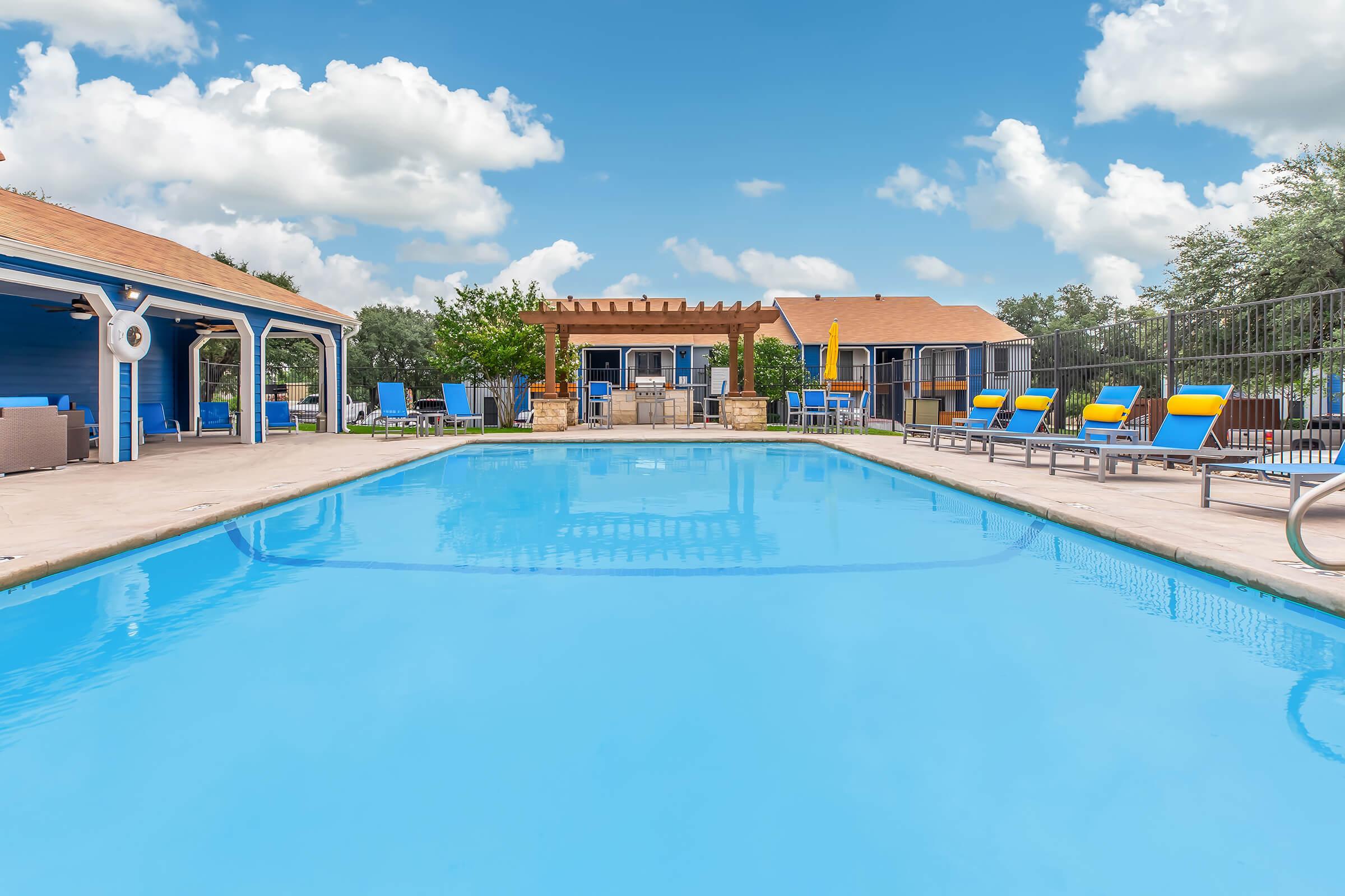 A clear blue swimming pool surrounded by lounge chairs. In the background, there are buildings with a pergola and seating area, under a bright blue sky with fluffy clouds. The scene conveys a relaxing outdoor atmosphere, perfect for leisure and enjoyment.