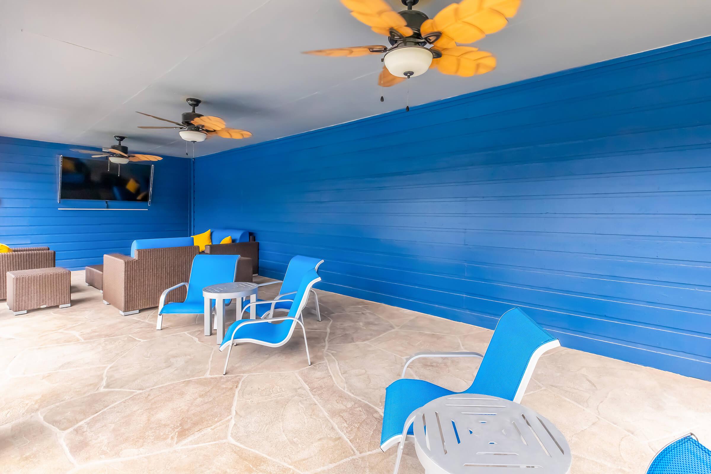 A modern outdoor seating area featuring a blue wall and light stone flooring. There are two ceiling fans with yellow blades, a wall-mounted TV, and various furniture pieces, including brown wicker chairs and a blue table with white metal chairs, creating a comfortable and inviting space.