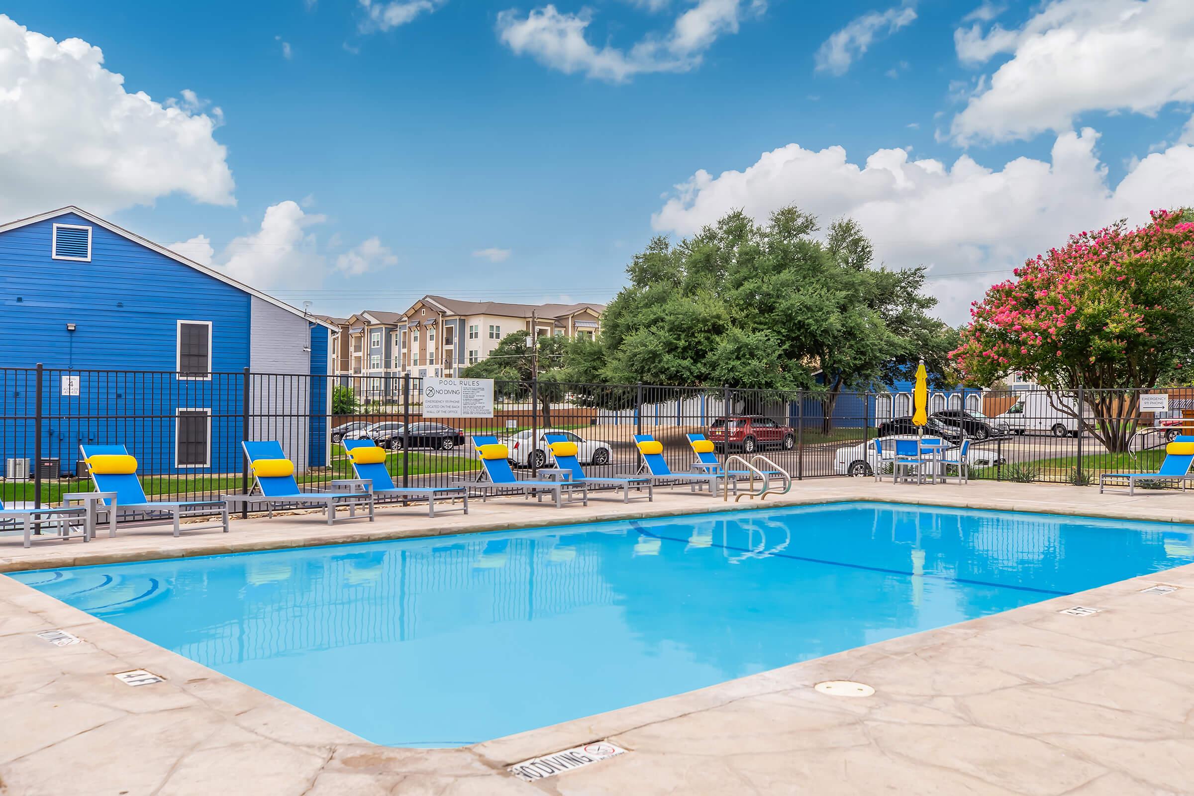 A swimming pool with clear blue water surrounded by yellow and gray lounge chairs. In the background, there's a blue building and several trees. The sky is bright with scattered clouds, and there are vehicles parked nearby. The area appears well-maintained and inviting for relaxation.