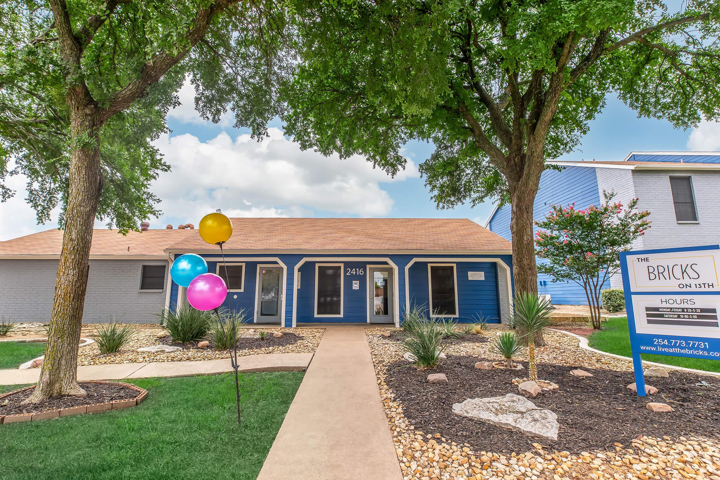 A charming multi-unit building with a blue exterior and tan roof, featuring landscaped gardens with rocks and shrubs. The entrance has a welcoming path leading to the door, and colorful balloons are tied to a nearby railing. A sign displays "The Bricks on 5th," indicating office hours and contact information.