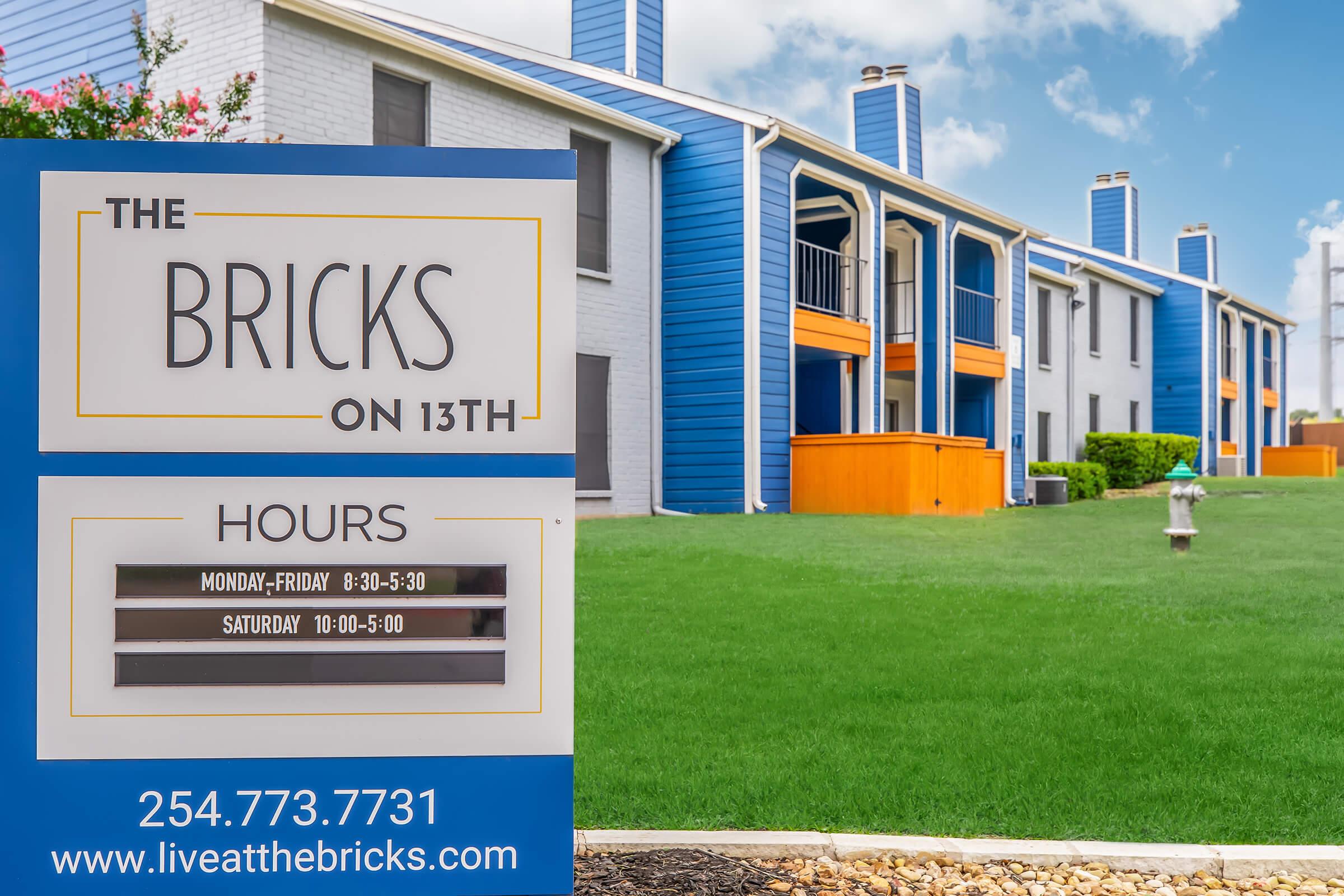 Sign for "The Bricks on 13th" apartment complex featuring hours of operation: Monday-Friday 8:30 AM - 5:30 PM, Saturday 10:00 AM - 5:00 PM. In the background, there are blue and white apartments with a green lawn and blue skies. Contact number displayed at the bottom.