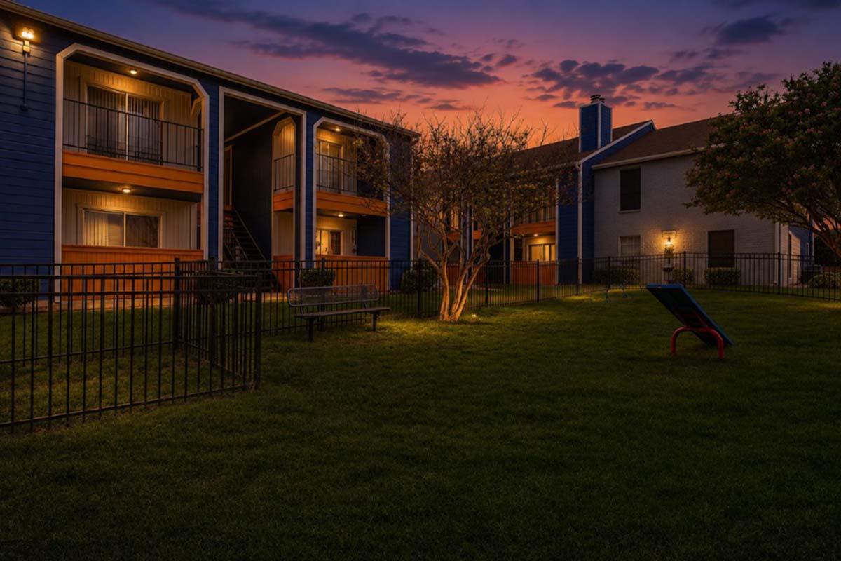 An evening scene of an apartment complex featuring two buildings with balconies and lit pathways. A small tree is illuminated in the foreground, surrounded by green grass. The sky showcases a gradient of colors at sunset, creating a serene atmosphere. A bench and lounge chair are visible in the lawn area.