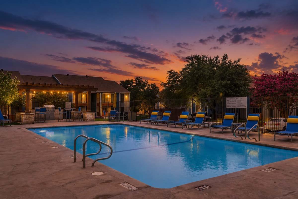 A tranquil outdoor swimming pool area at sunset, featuring a clear blue pool surrounded by lounge chairs. Lush trees and flowering plants decorate the space, with warm light emanating from nearby buildings, creating a serene and inviting atmosphere.