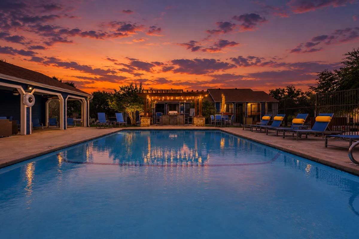 A serene swimming pool at dusk, reflecting the vibrant orange and purple sky. The pool area features lounge chairs and is surrounded by trees and buildings. Soft lighting enhances the inviting atmosphere, creating a tranquil setting for relaxation.