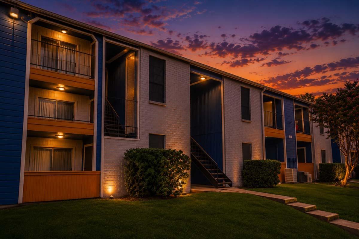 A modern apartment building at dusk, featuring a mix of blue and white exteriors. The scene shows balconies illuminated by warm lighting, lush green grass, and a colorful sky with clouds illuminated by sunset hues. Pathway steps lead to the entrance, creating a welcoming atmosphere.