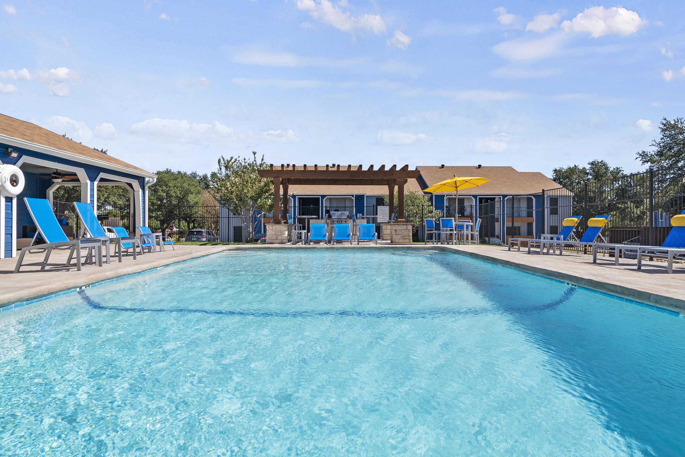 A spacious outdoor swimming pool with clear blue water, surrounded by lounge chairs in vibrant colors. In the background, there's a pergola and a building with large windows. Bright yellow umbrellas provide shade, and the area is landscaped with trees. The sky is mostly clear, creating a sunny atmosphere.