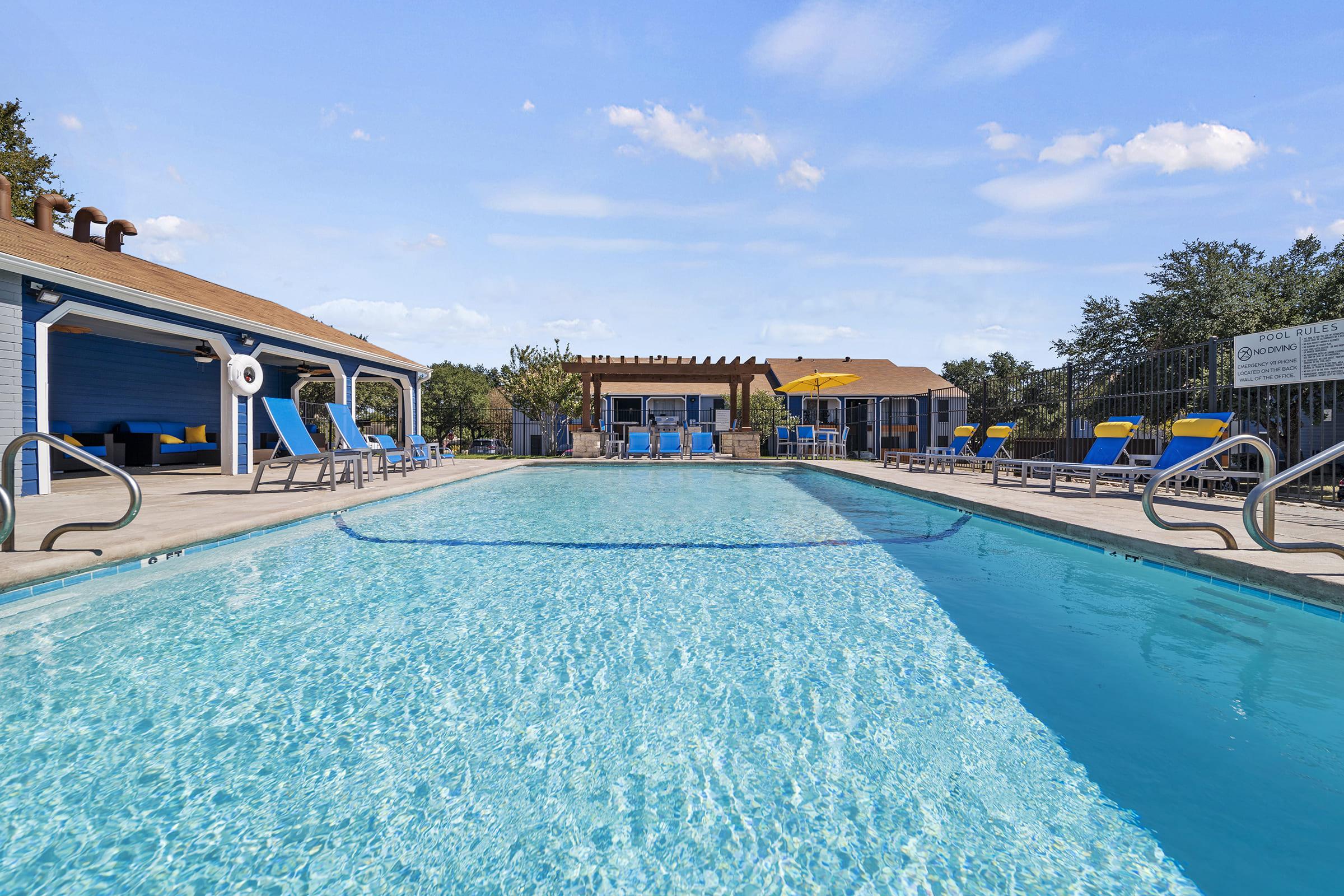 A clear swimming pool surrounded by lounge chairs with blue and yellow cushions. In the background, there is a recreational area with shaded seating and trees. The sky is partly cloudy, creating a bright and inviting atmosphere.